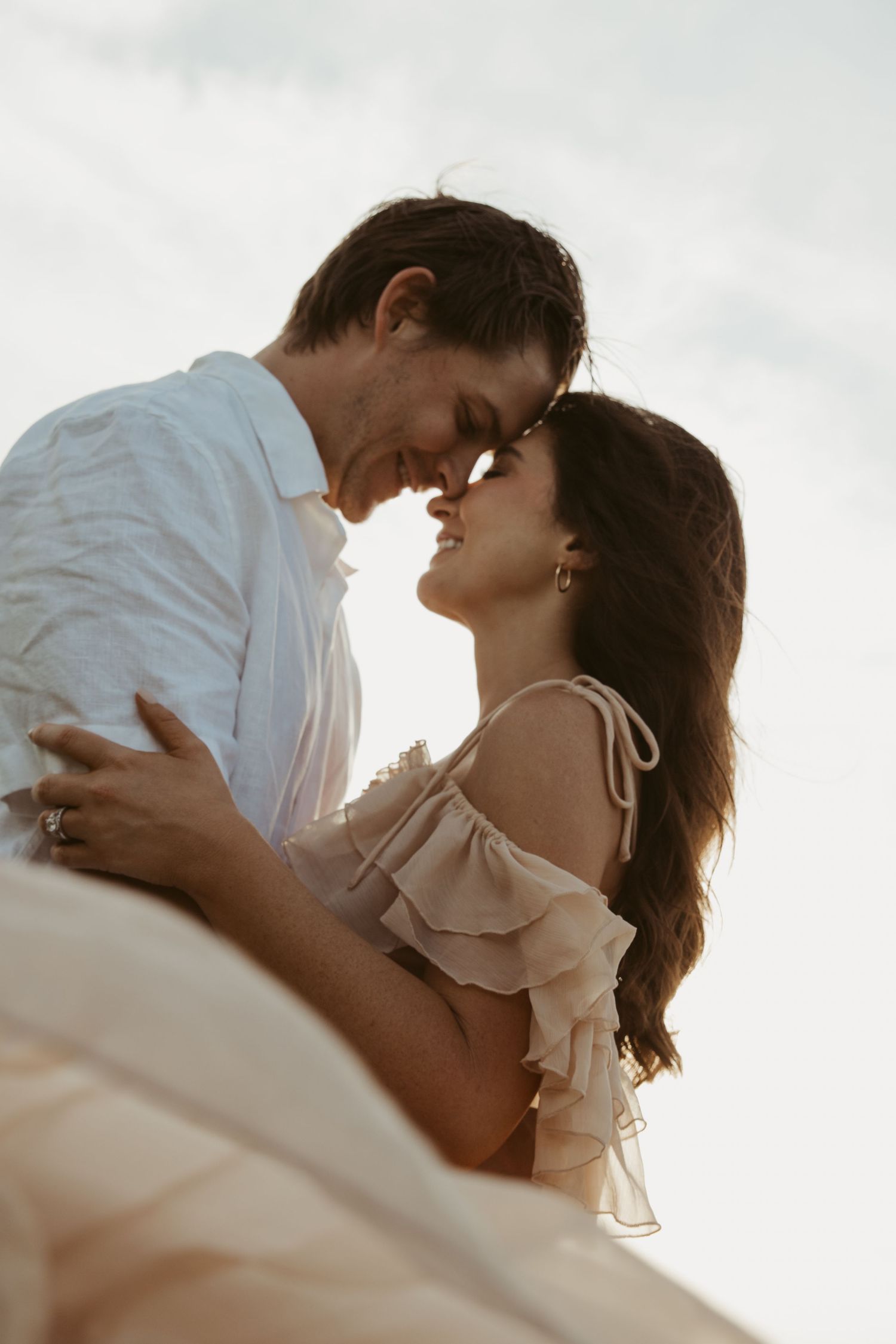 A romantic couple embraces in soft sunlight wearing white and beige outfits during an intimate outdoor moment.