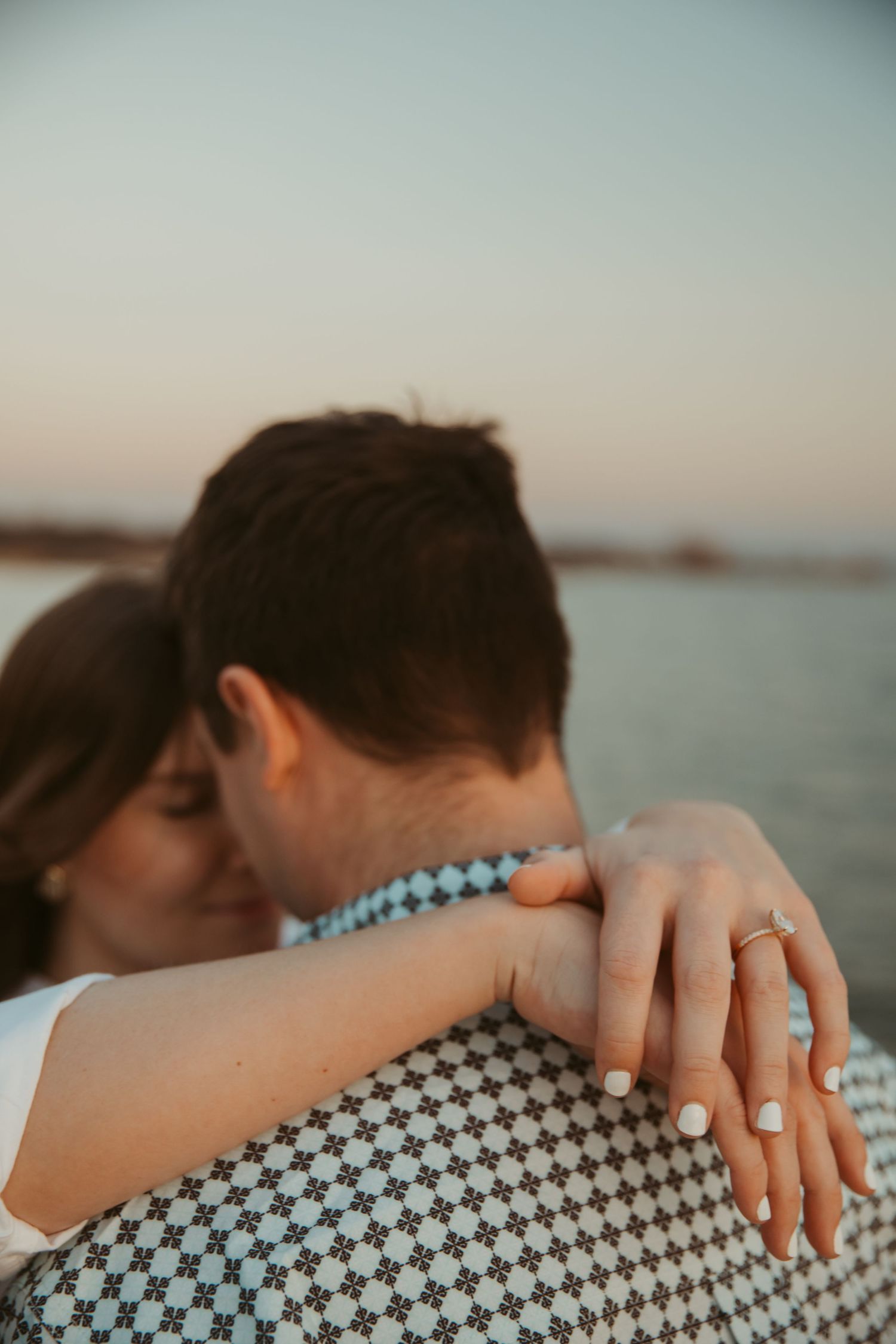 Close-up of woman's hand with engagement ring resting on man's shoulder as couple embraces near water at sunset.