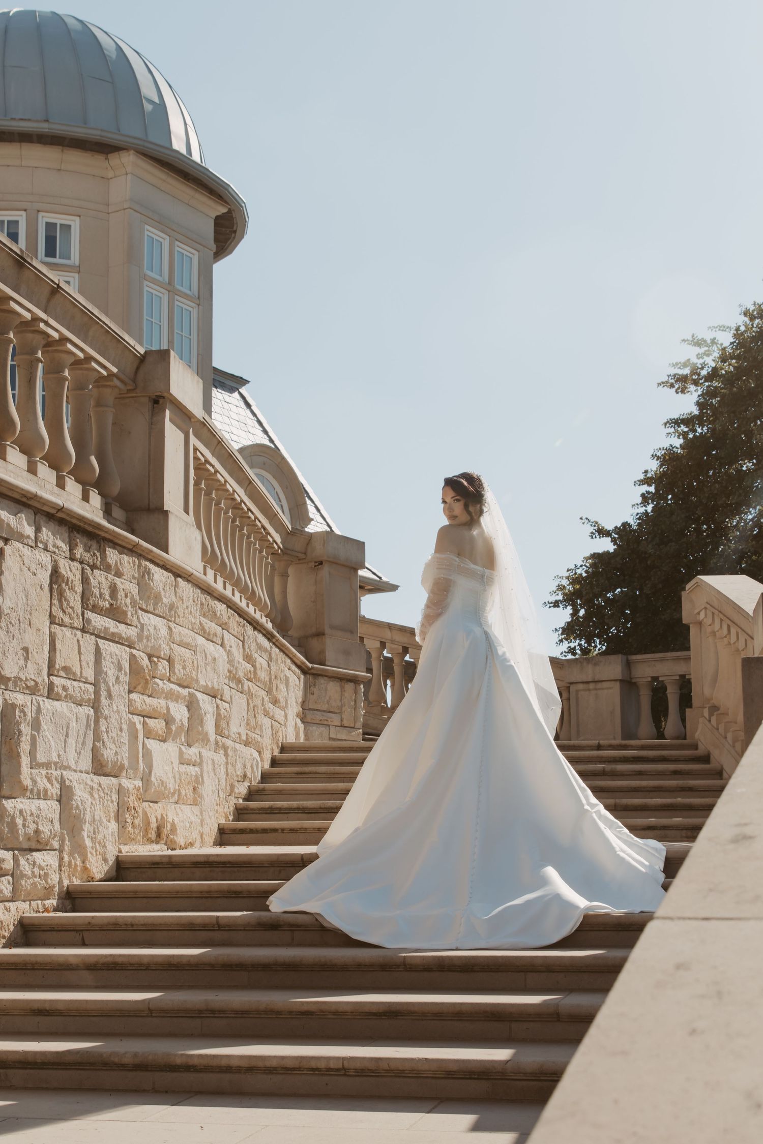 A white wedding dress flows elegantly as someone descends grand stone stairs at a luxurious venue.
