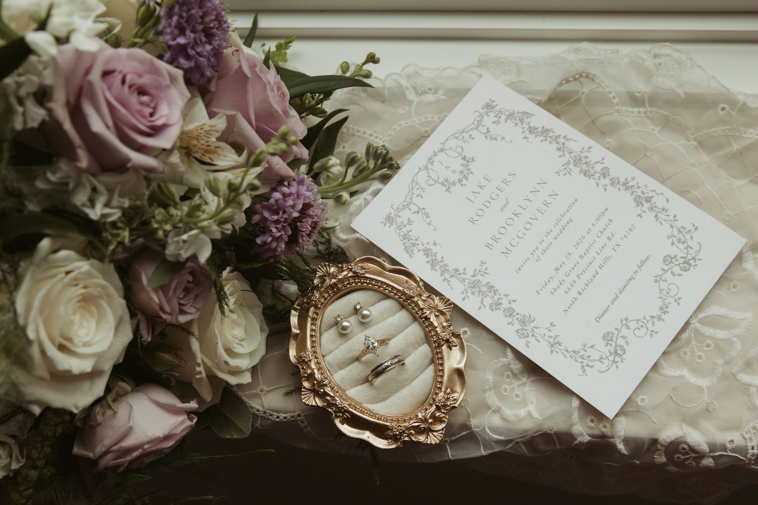 Wedding flat lay with purple roses, gold ring dish, jewelry, and elegant invitation on lace fabric.