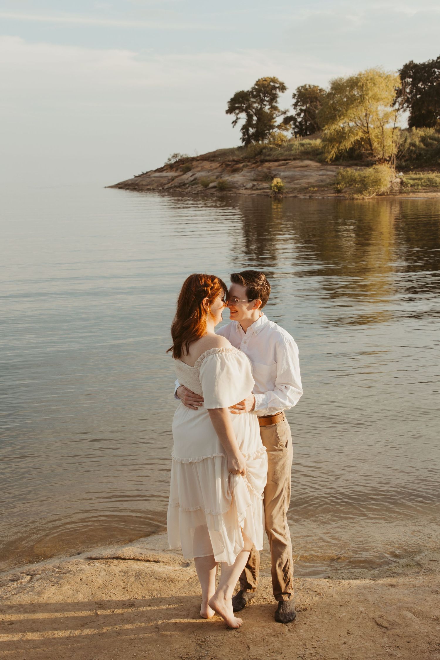 A romantic couple in white outfits shares an intimate moment on a rocky beach at sunset.