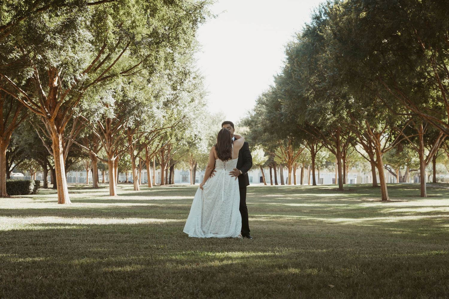 A couple in wedding attire poses together in a scenic tree-lined park setting during golden hour. Wedding Photographer DFW