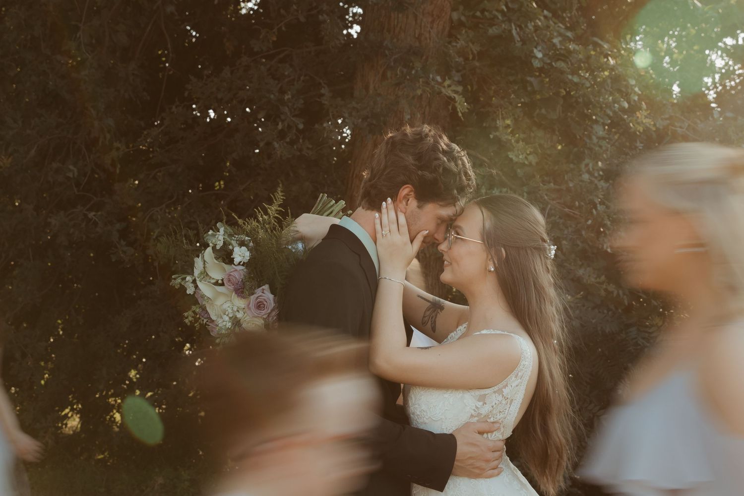 A bride and groom share a romantic kiss surrounded by lush greenery and soft golden light at their outdoor wedding.
