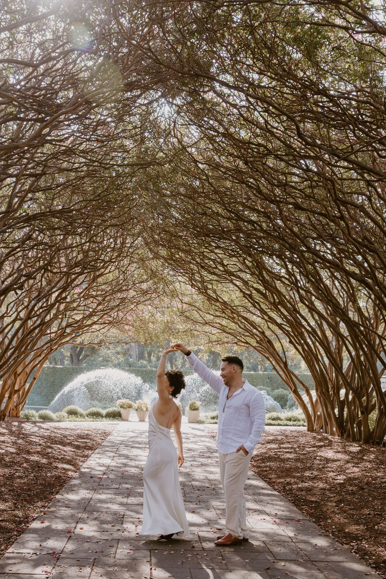 A romantic wedding photo under a natural archway of intertwined tree branches in a garden setting.