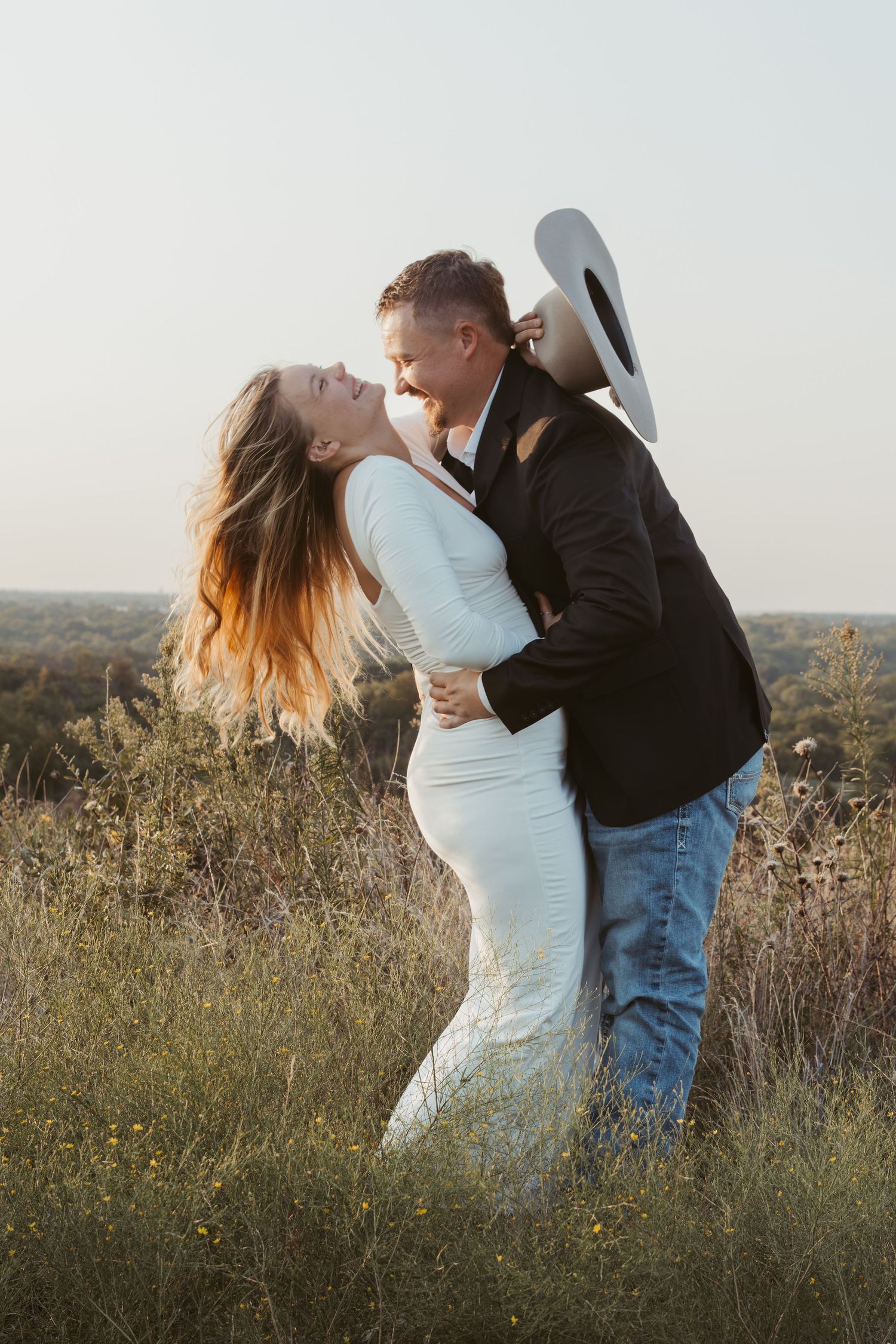 A couple shares a romantic embrace in a golden field at sunset while posing for engagement photos.