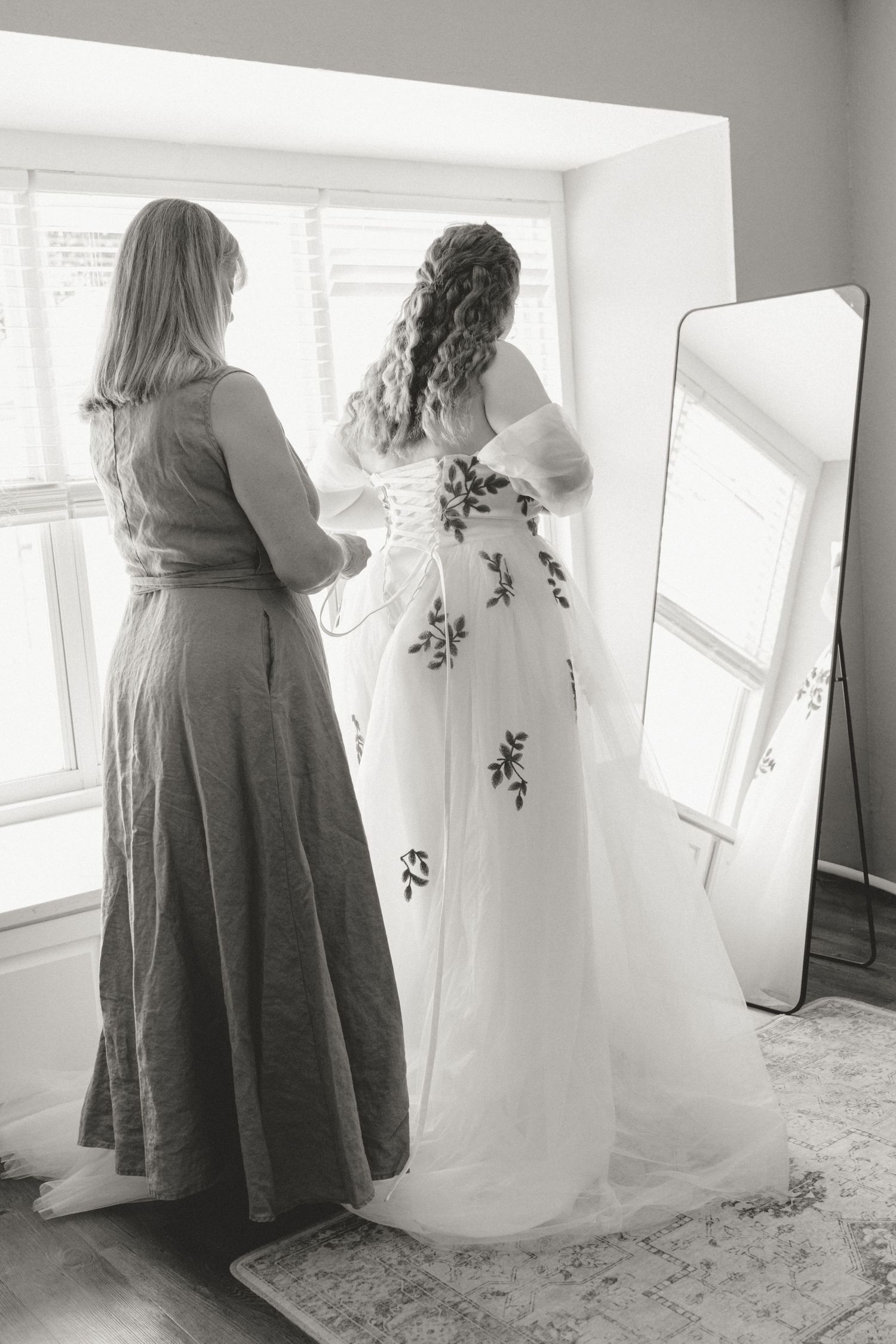 Two people admire a floral wedding dress near a window in black and white.