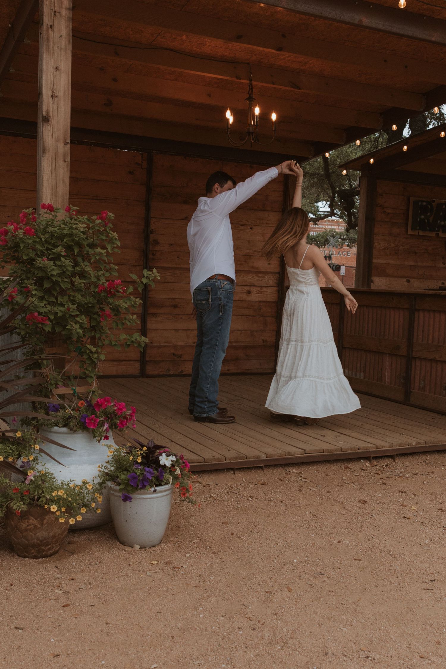 A couple dancing on a wooden deck decorated with colorful potted flowers and string lights overhead.