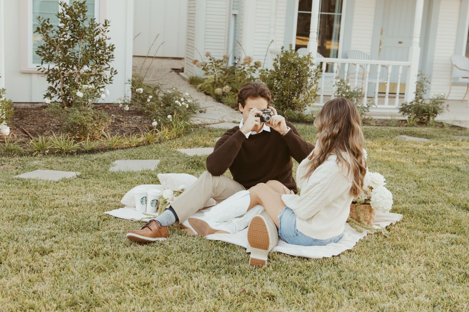 Two people relax on a picnic blanket in front of a white house with a porch on a sunny day.