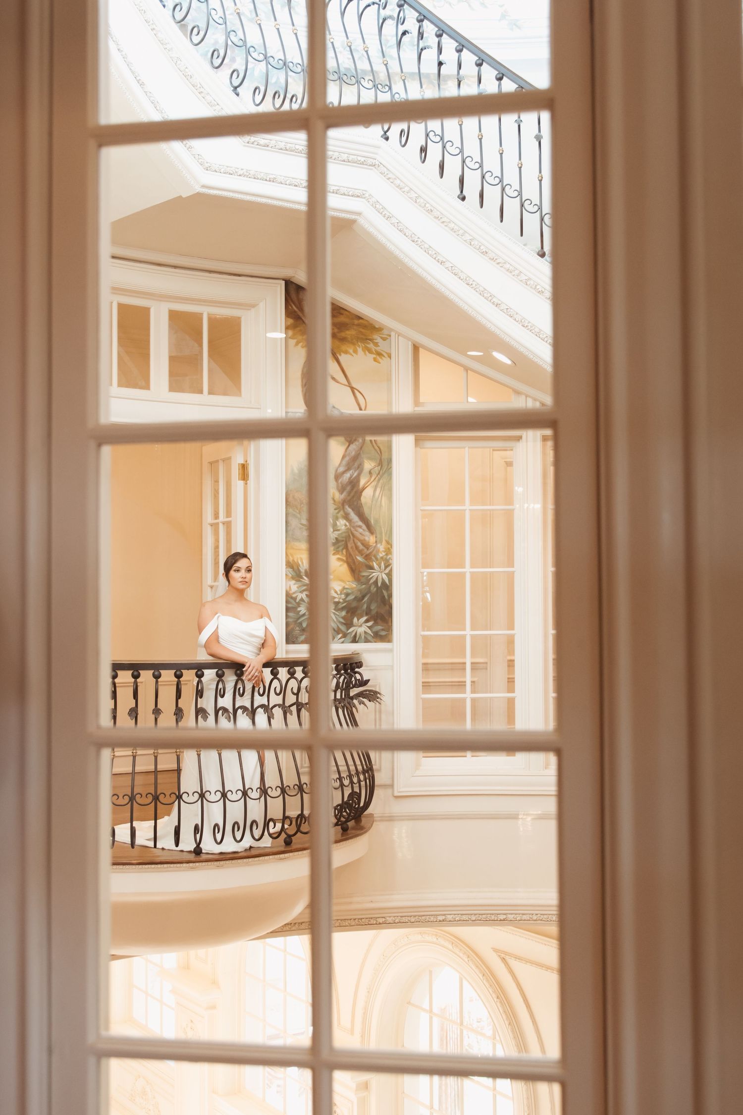 Elegant bride in white dress standing on ornate balcony of luxurious mansion, viewed through French doors.