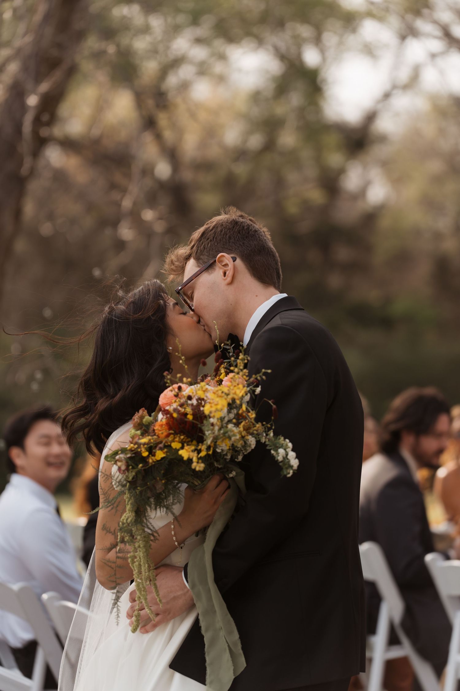 Bride and groom share a first kiss at outdoor wedding ceremony, bride holding colorful wildflower bouquet.