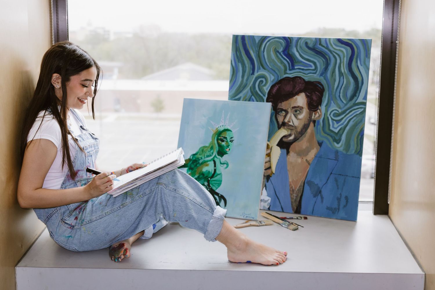 Artist sketches while sitting barefoot on floor next to colorful paintings featuring a green figure and blue portrait.