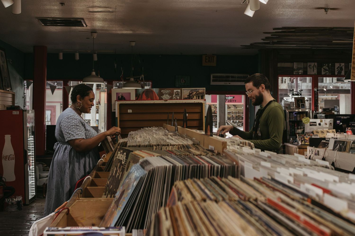 People browse through vinyl records in a dimly lit vintage record store with rows of albums in cardboard boxes.