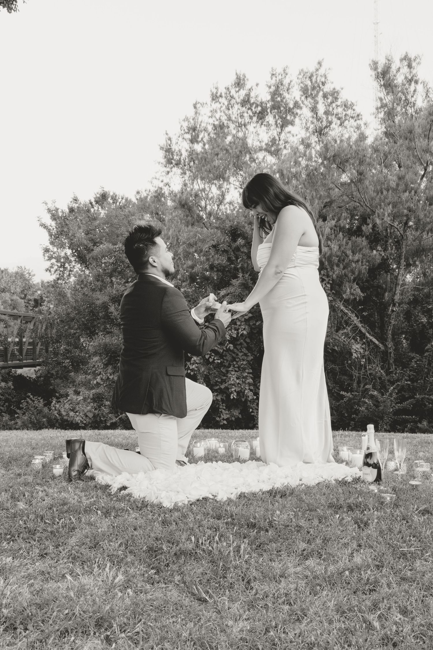 Black and white proposal photograph taken outdoors with wine bottles and a picnic blanket on grass near trees.