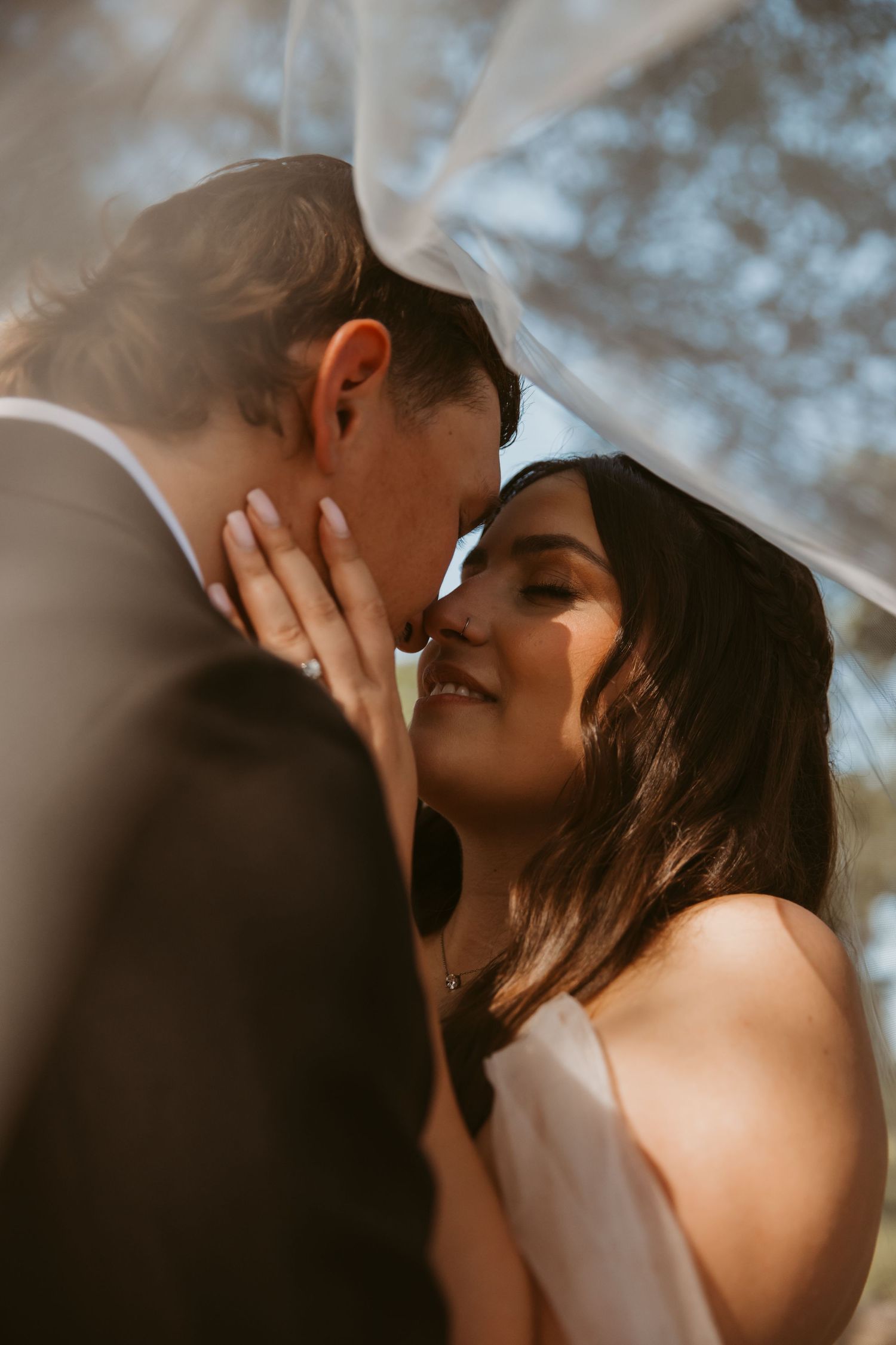 A bride and groom share a romantic moment under a wedding veil, their foreheads touching in golden sunlight.