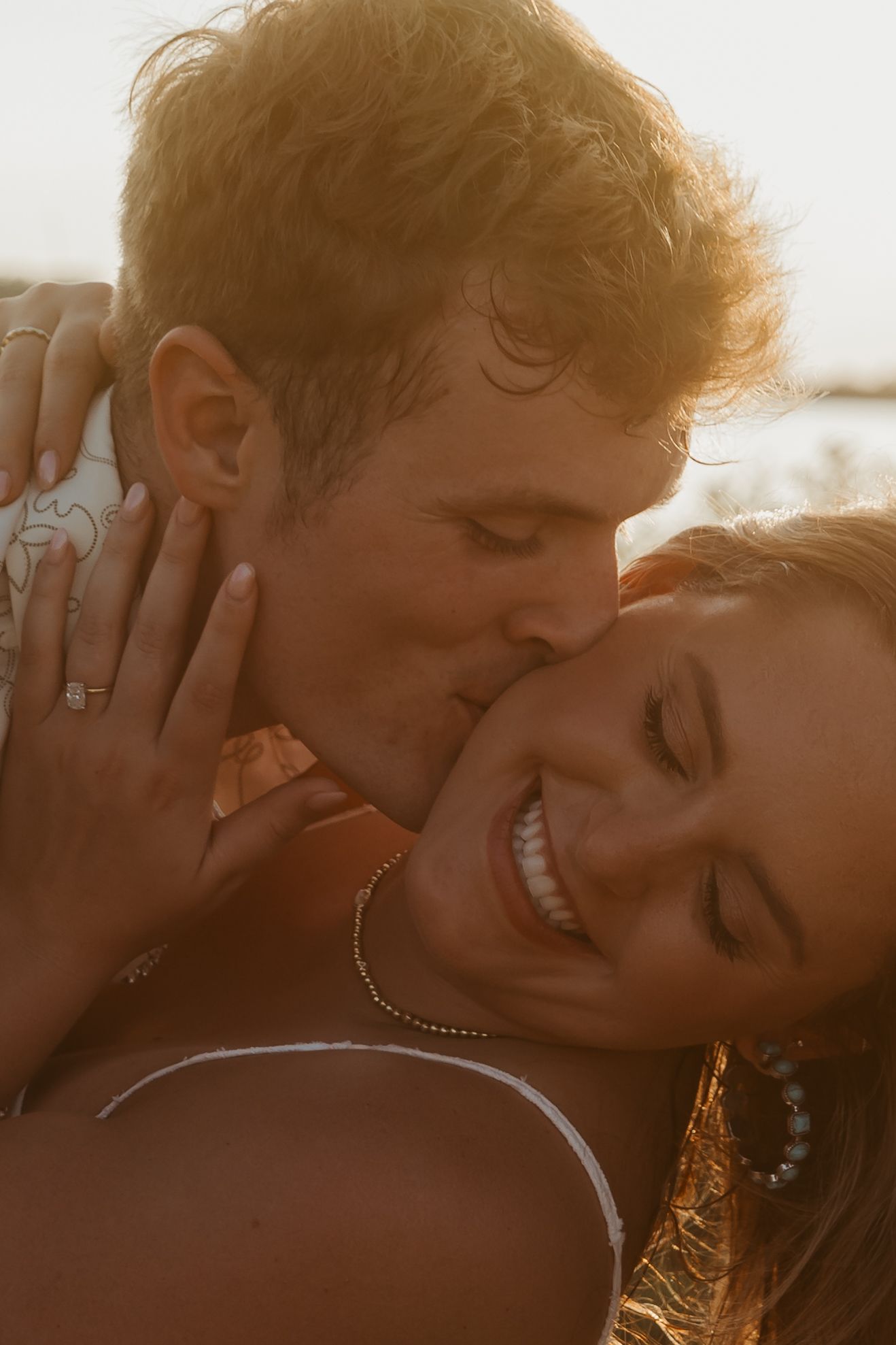 A man with red hair kisses a smiling woman on the cheek during a golden sunset outdoor moment.
