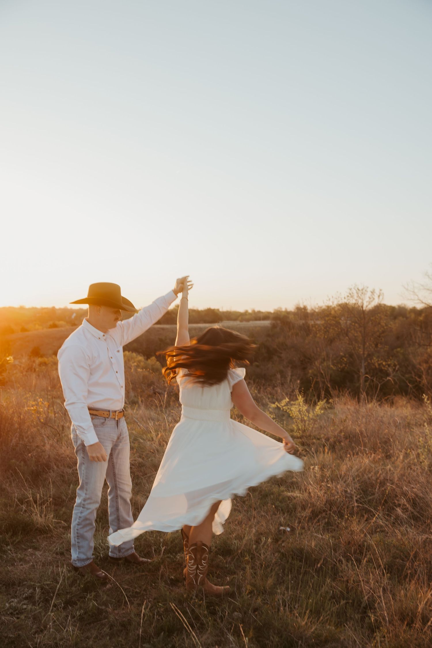 Couple dancing at golden hour in a field, woman in white dress and cowboy hat being twirled by man in white shirt.