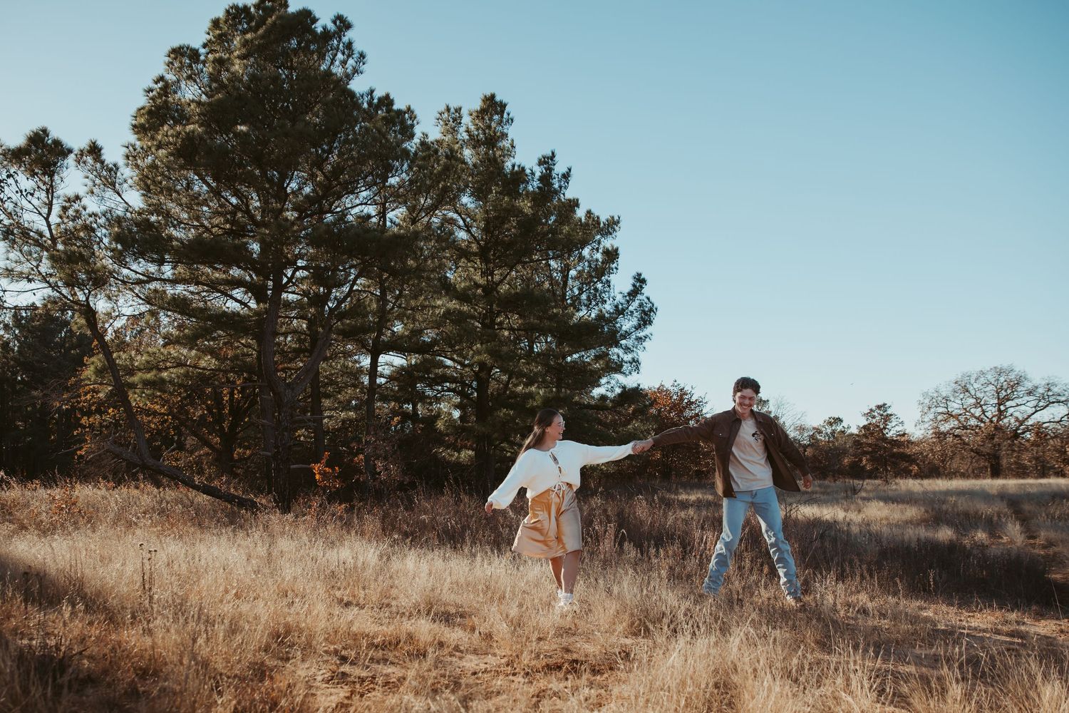 A couple walks through a golden field at sunset with tall pine trees in the background during a romantic photoshoot. engagement photos DFW