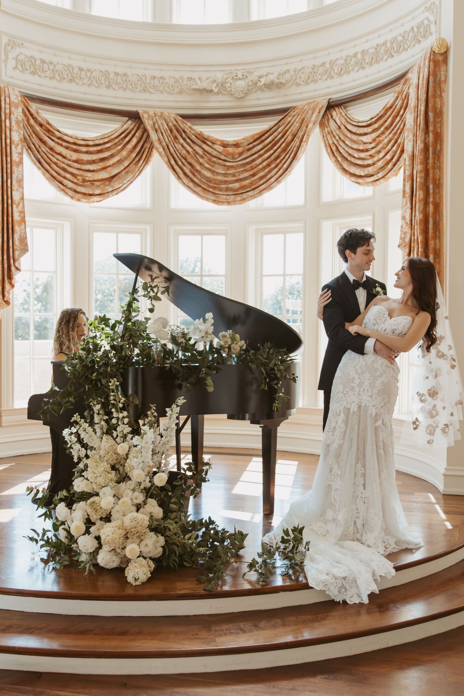 Elegant ballroom wedding scene with grand piano decorated in white florals and draped windows.