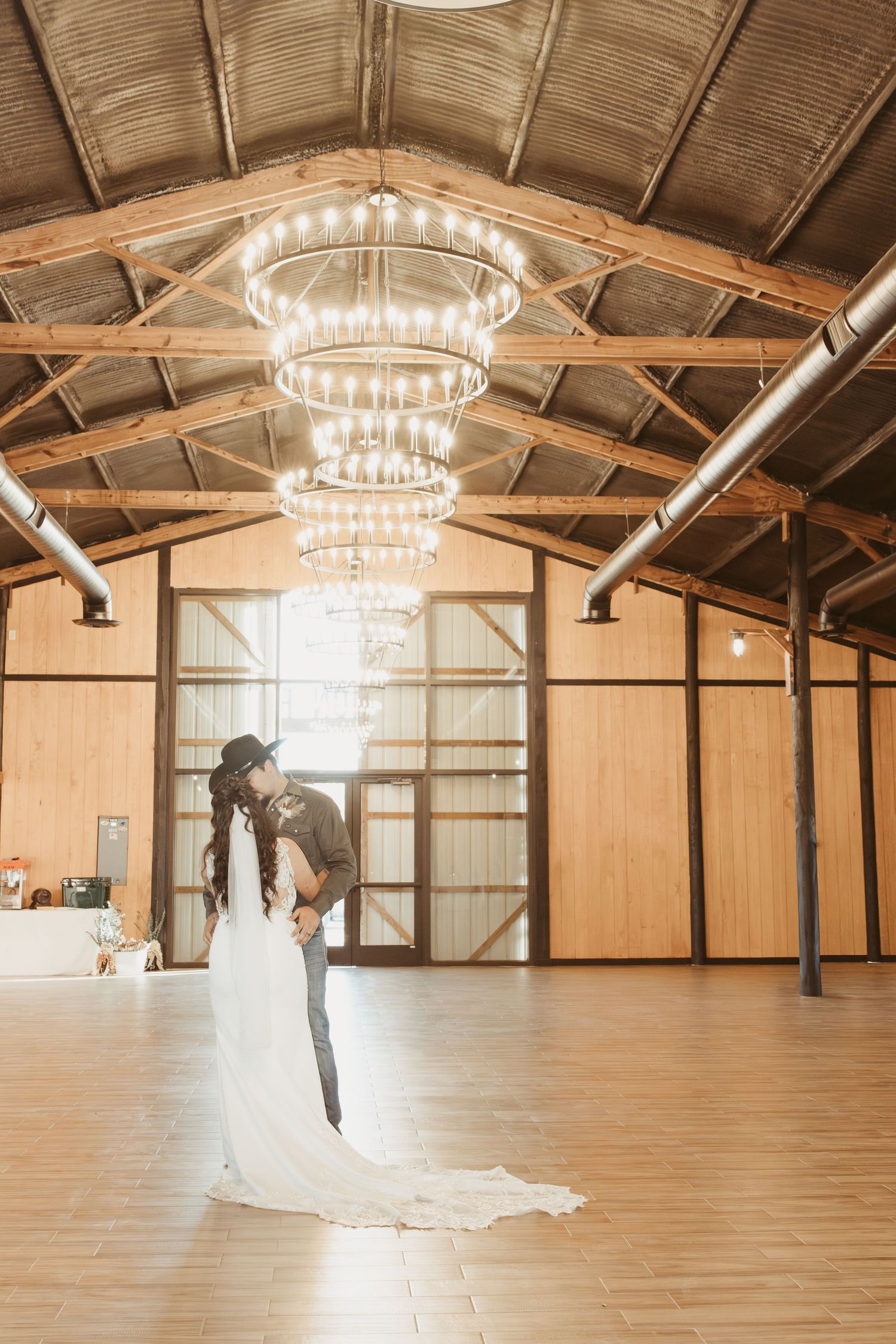 A romantic moment captured in a rustic barn venue with tiered crystal chandeliers and wooden beam ceiling details. Wedding photographer DFW