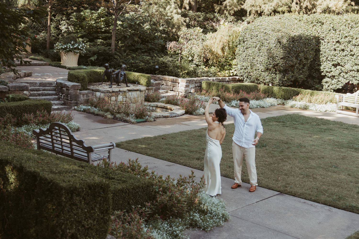 A couple shares a romantic moment on a sunny garden path surrounded by lush greenery and stone walls.