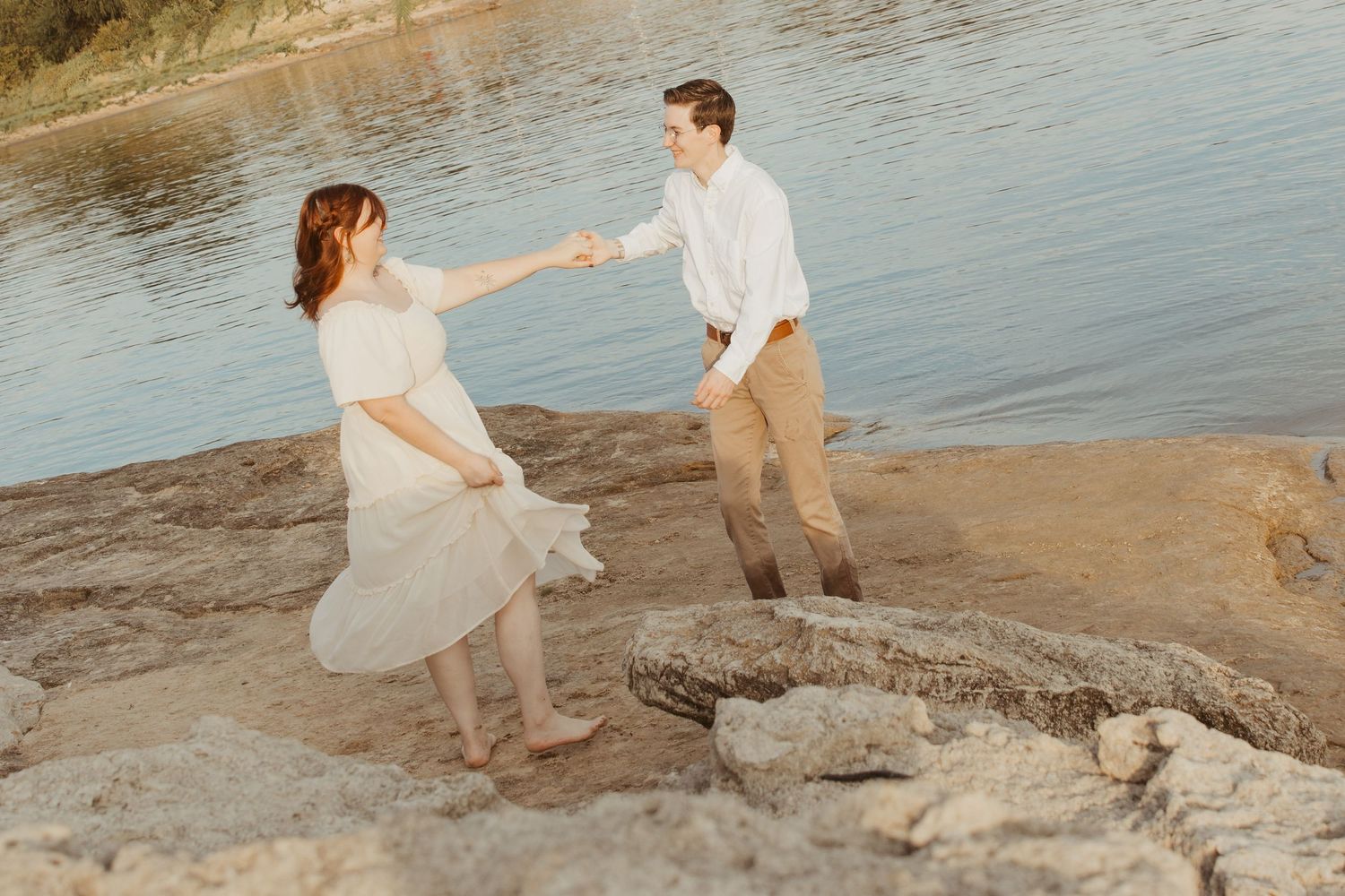 A couple in light colored clothing explores the rocky shoreline together.