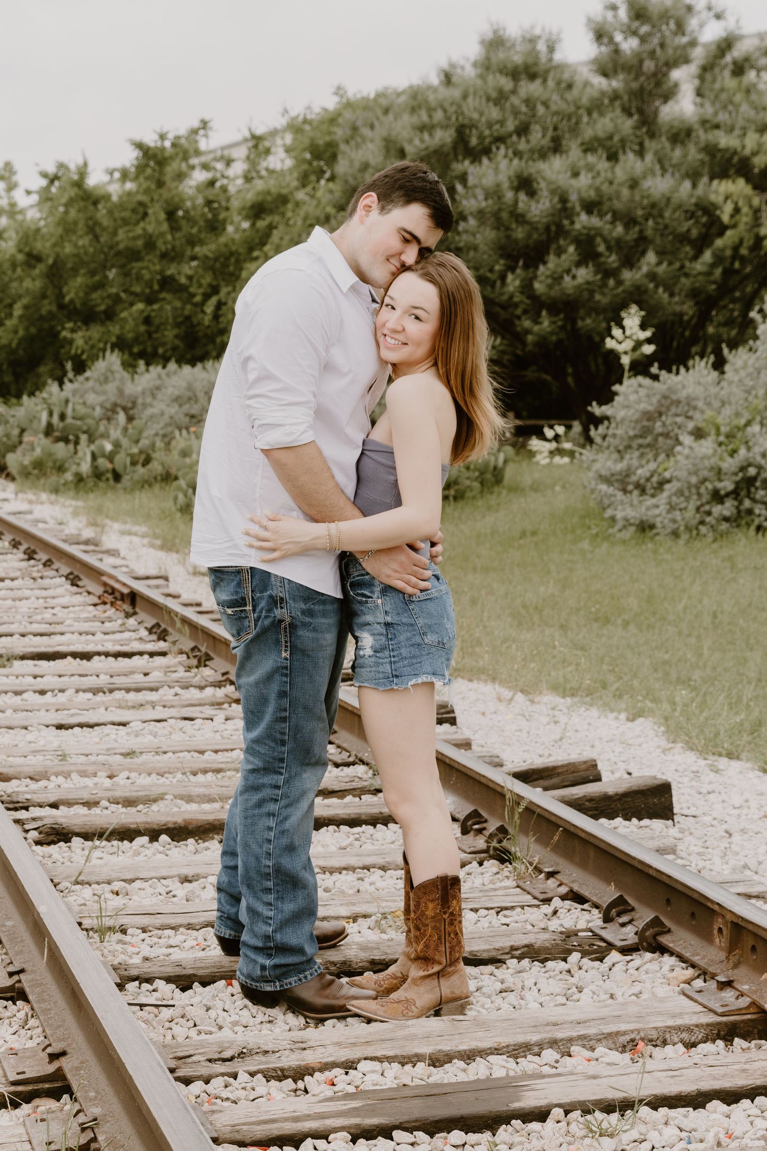 A romantic couple embraces on rustic train tracks surrounded by greenery on a sunny summer day.