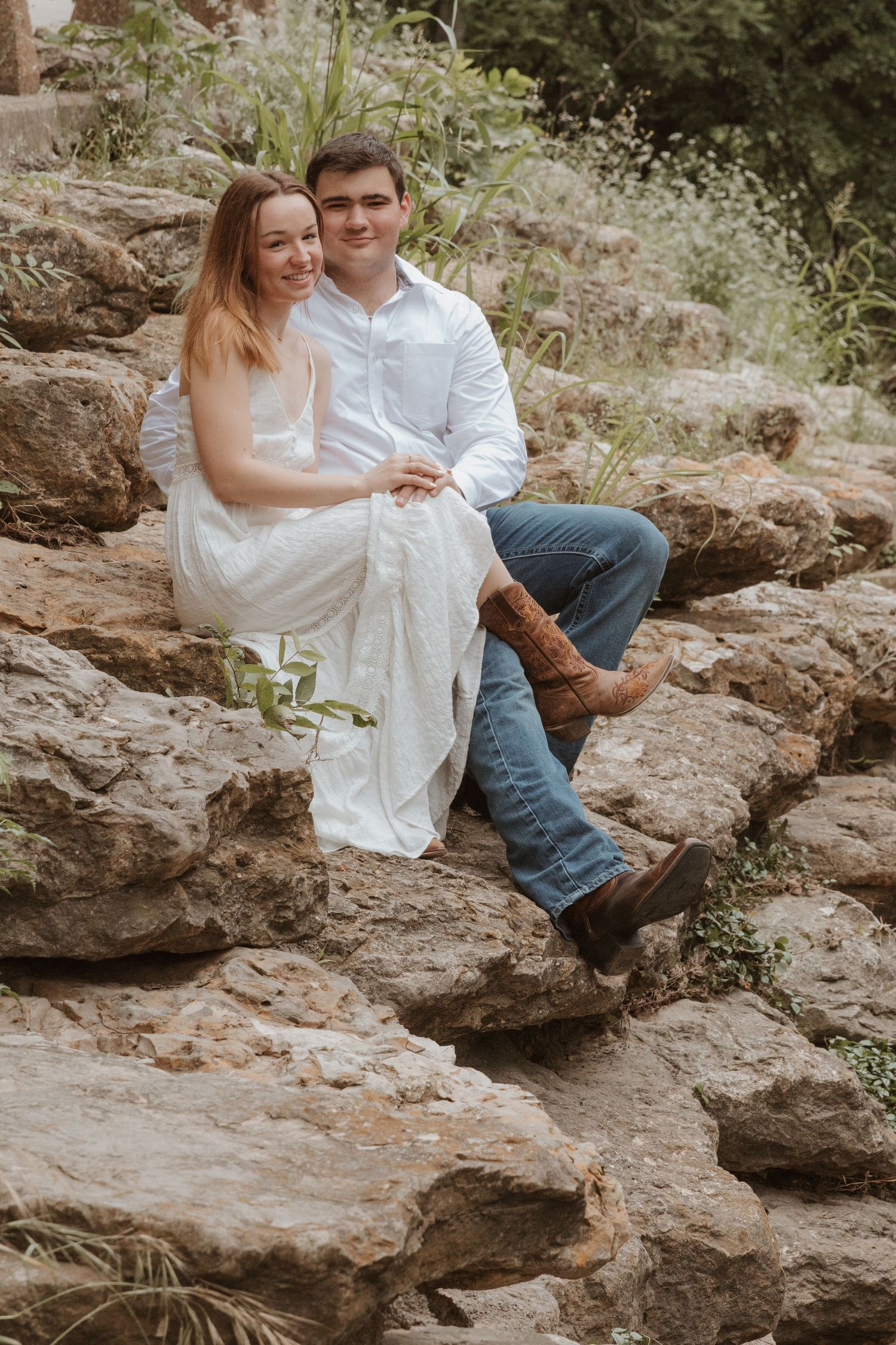 A couple in white and jeans sits together on rocky terrain during a romantic outdoor photo session.