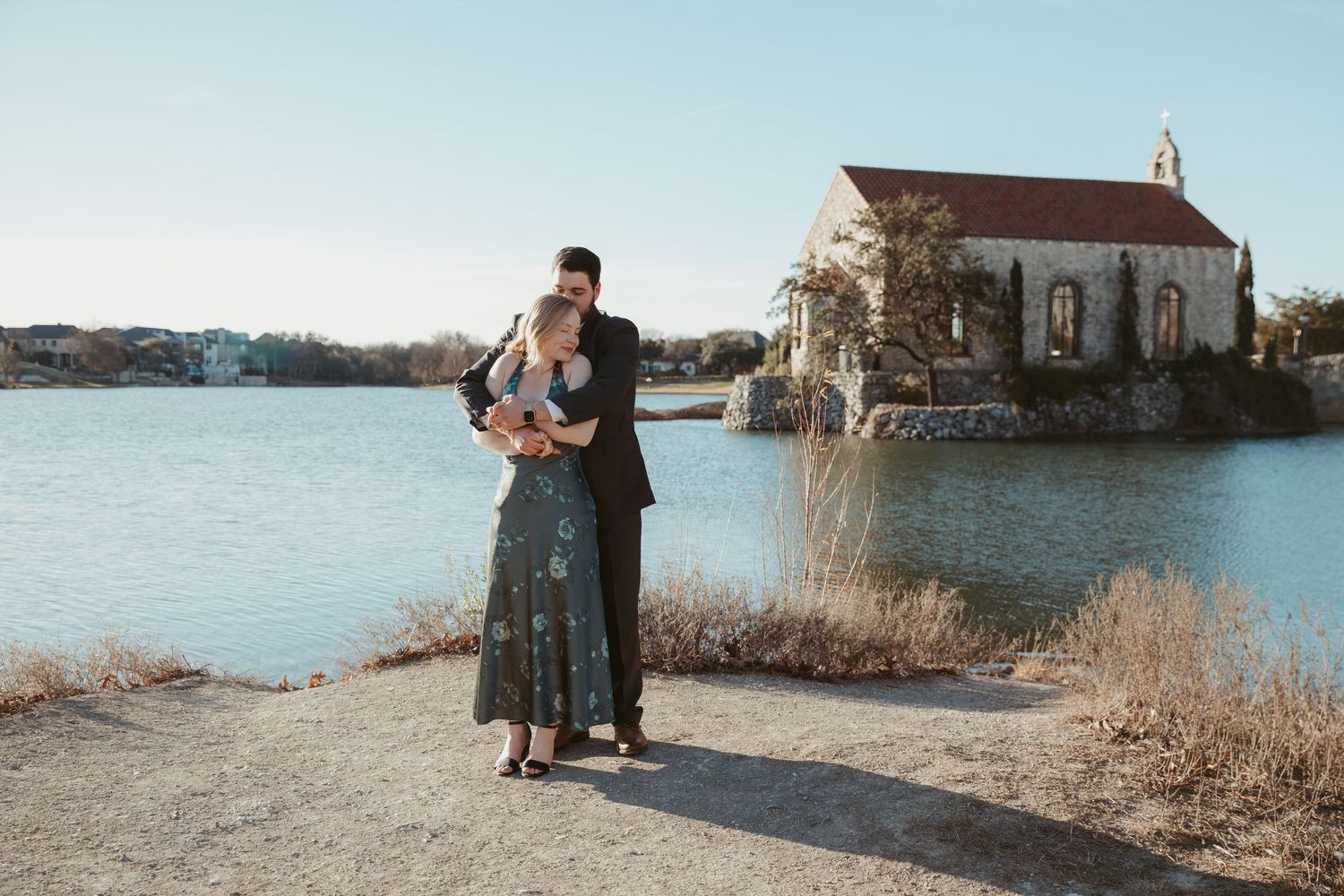 Wedding couple poses near a waterfront church during golden hour with scenic coastal views.