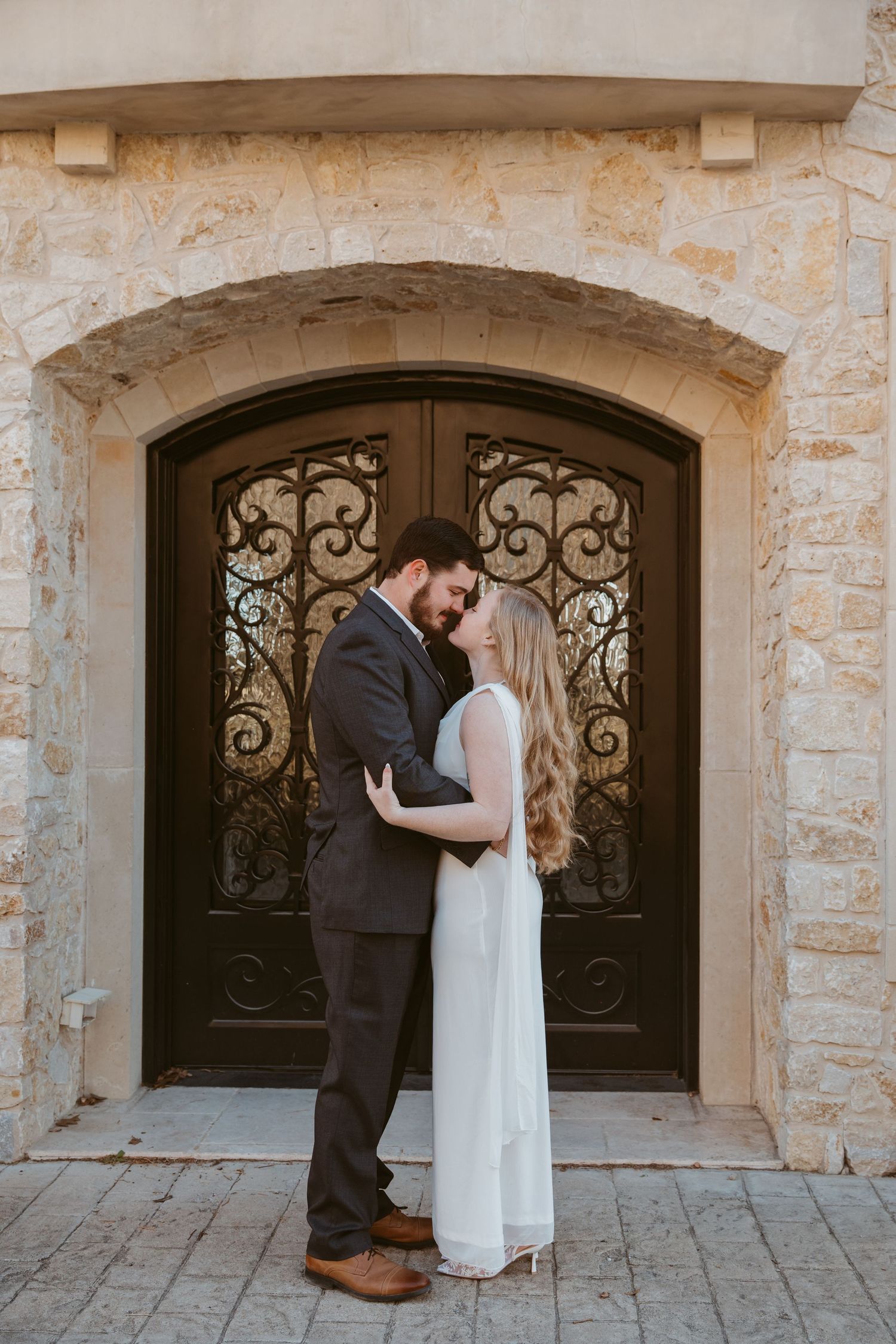A couple shares an intimate moment in front of an ornate wrought iron door at a stone building entrance.