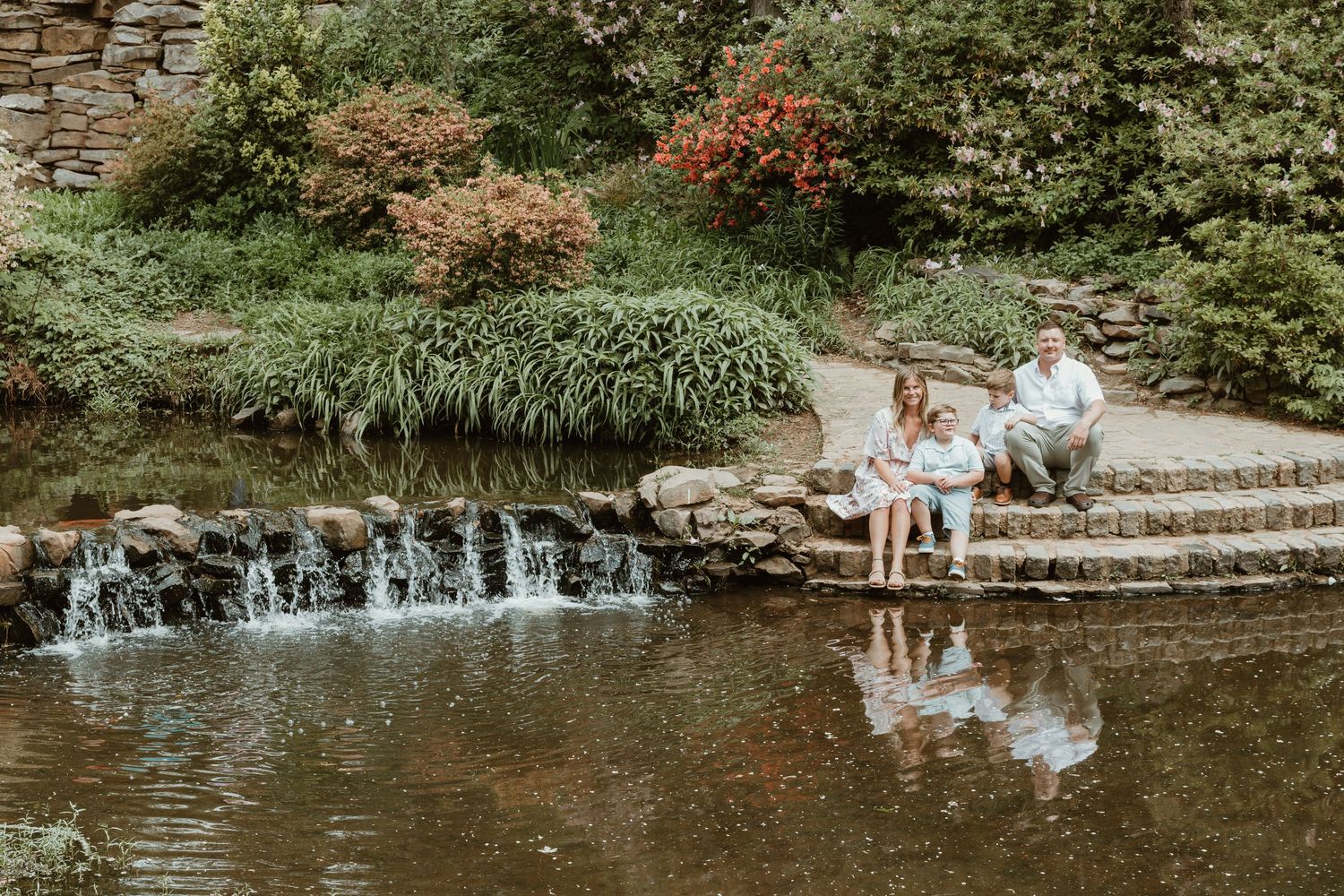 Family relax on stone steps by a peaceful pond with small waterfalls and flowering shrubs.
