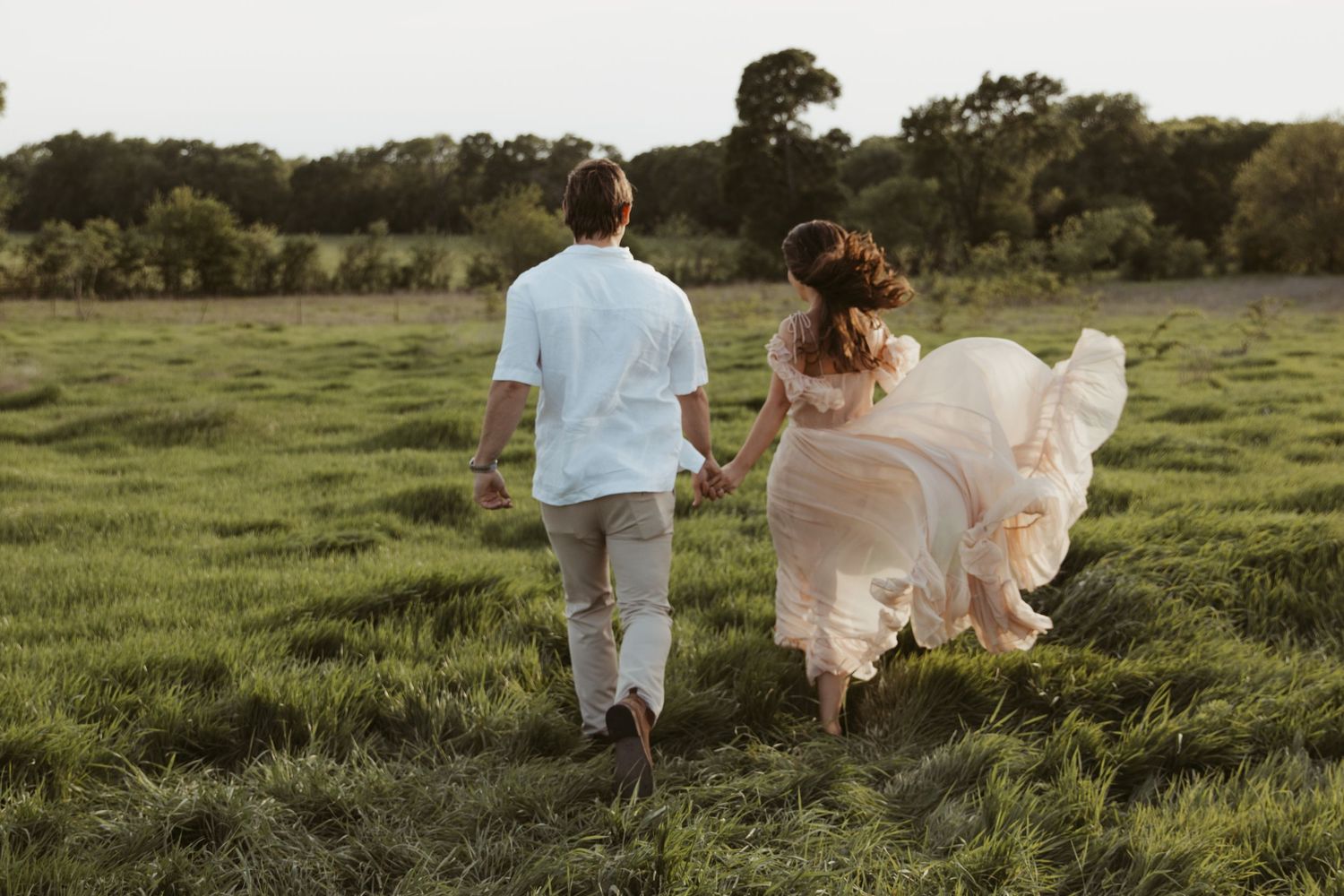 Two people run joyfully through a green meadow at sunset with one wearing a flowing dress that catches the breeze.