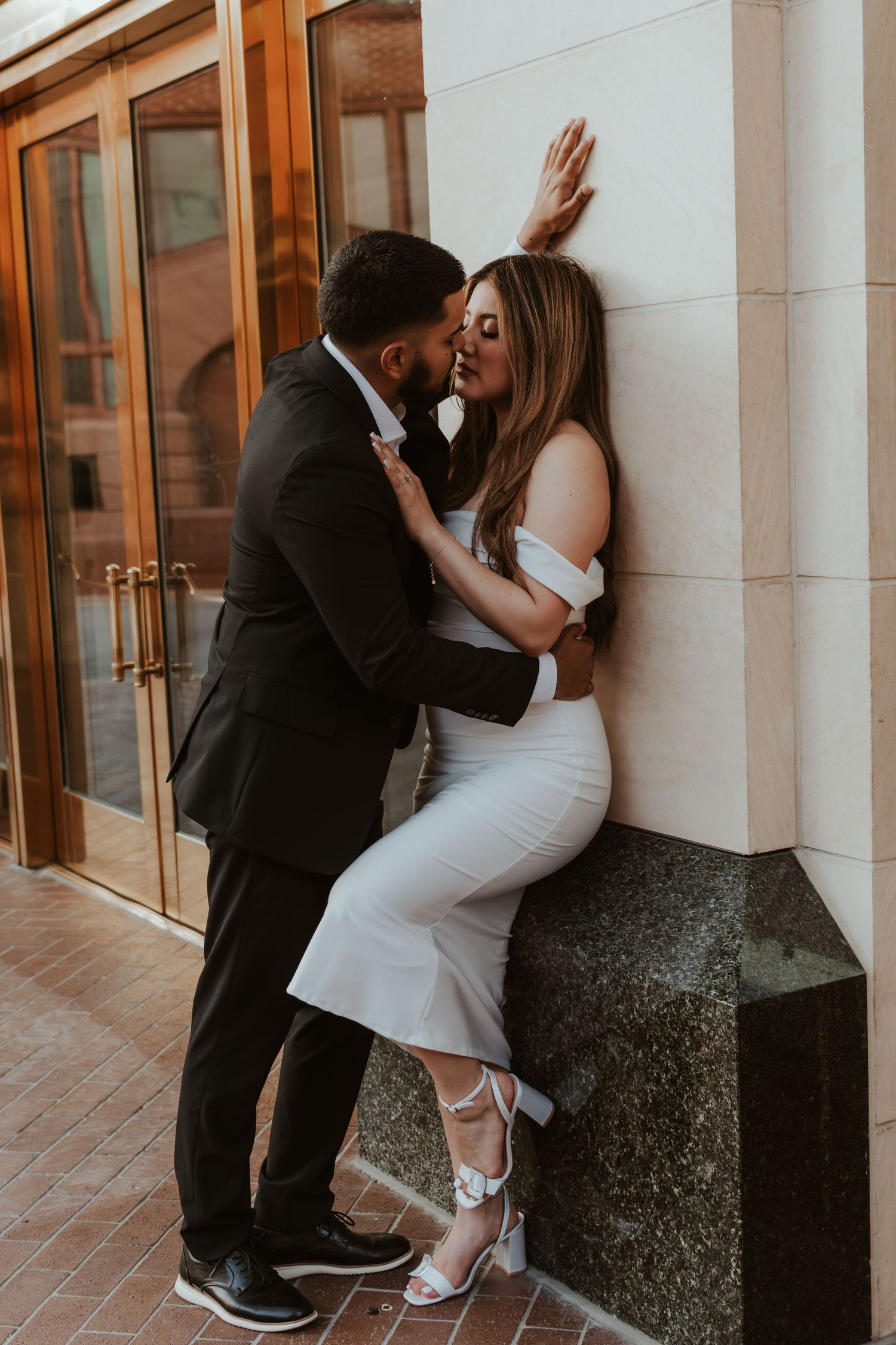 A romantic couple in formal attire sharing an intimate moment against a wall near glass doors.