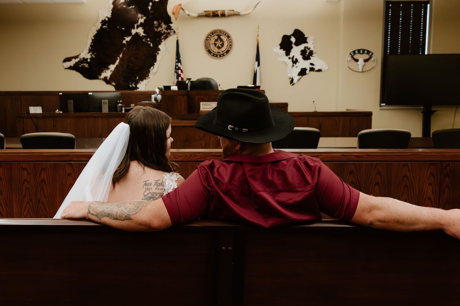 A back view of a couple sitting together in a courtroom, wearing a red t-shirt and white wedding dress.