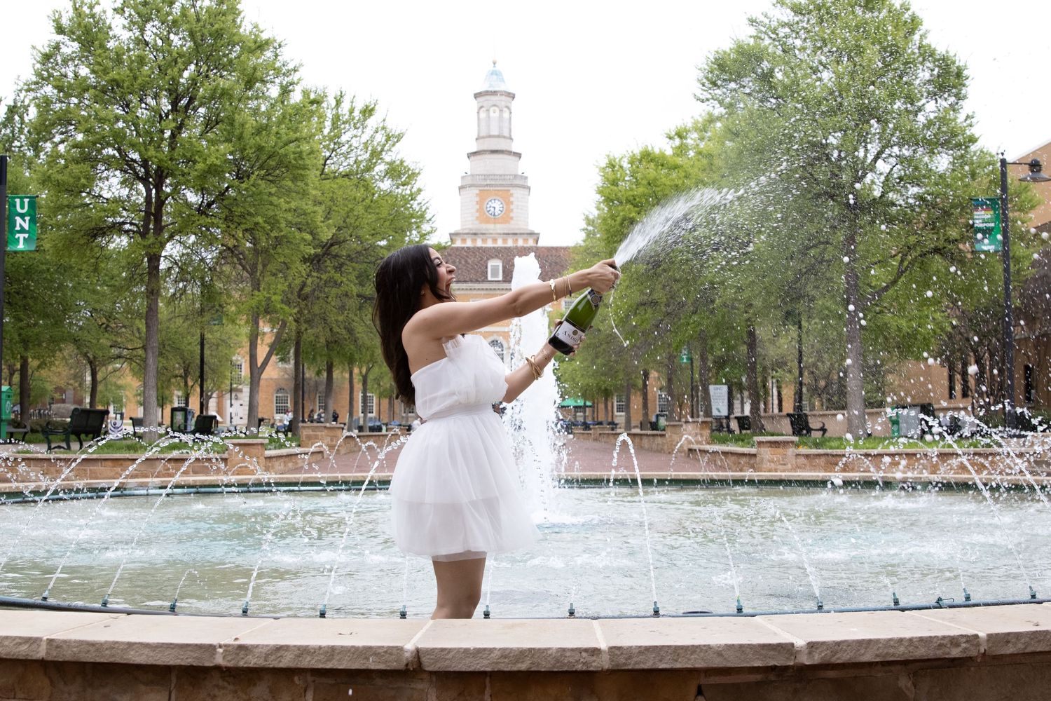 A person in a white dress stands by a fountain in front of a historic brick clock tower on a university campus.
