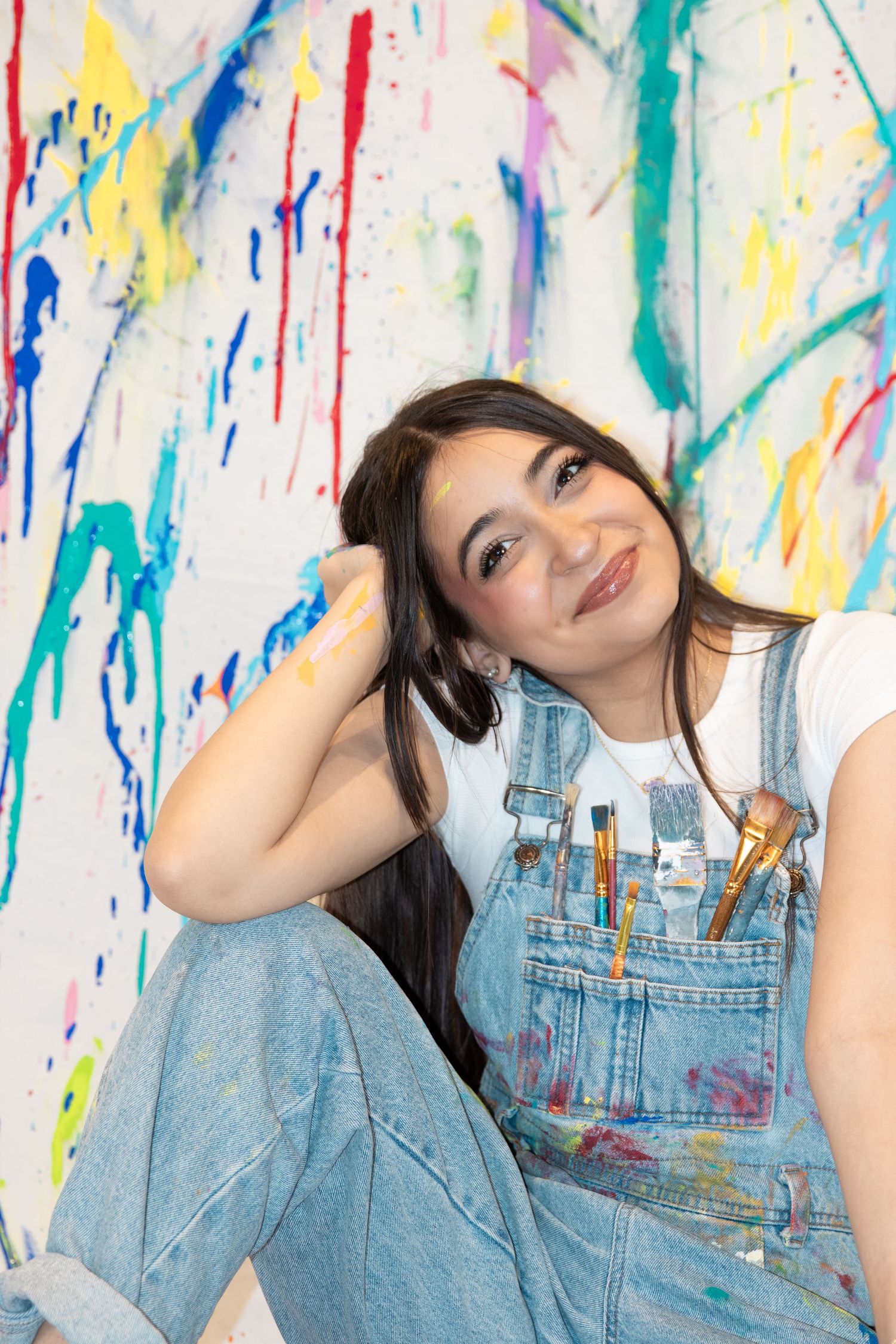 Artist in paint-splattered overalls sits against colorful drip-painted wall backdrop.