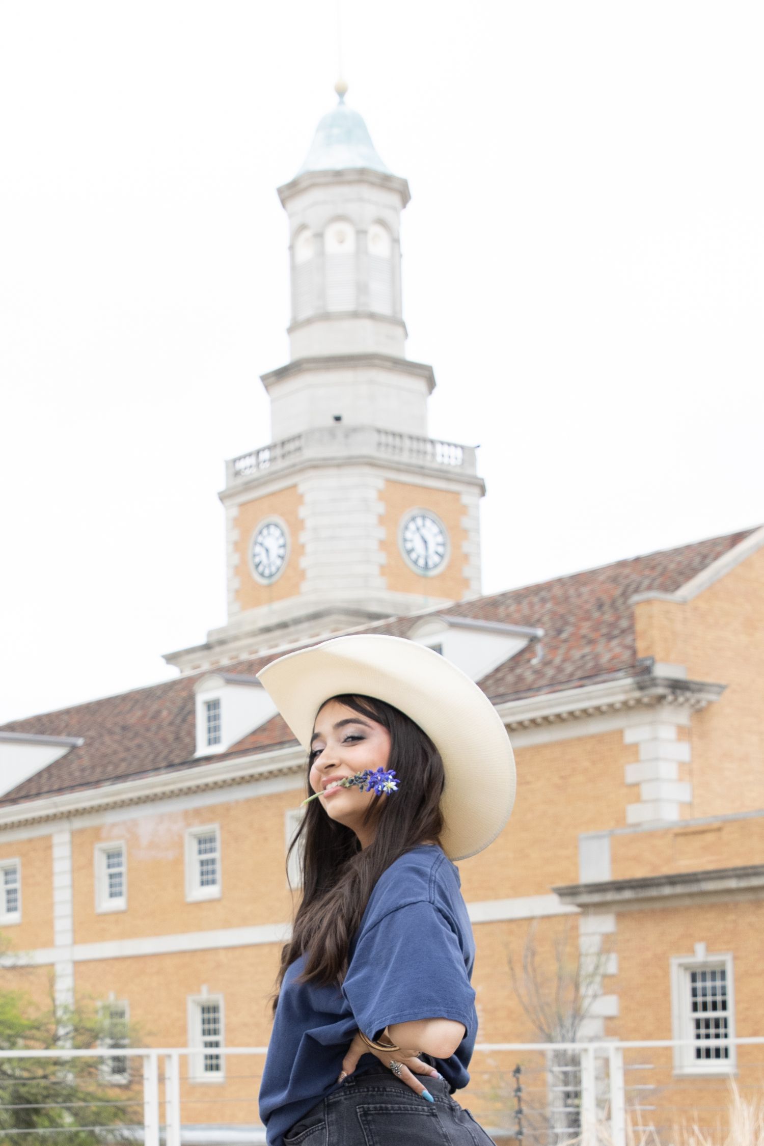 A person wearing a cowboy hat and navy shirt poses in front of a historic building with a white clock tower.