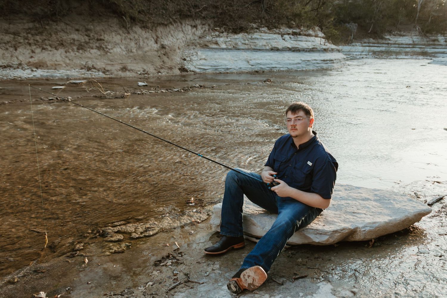 Someone relaxes while fishing on a fallen log at the edge of a river on a cloudy day.