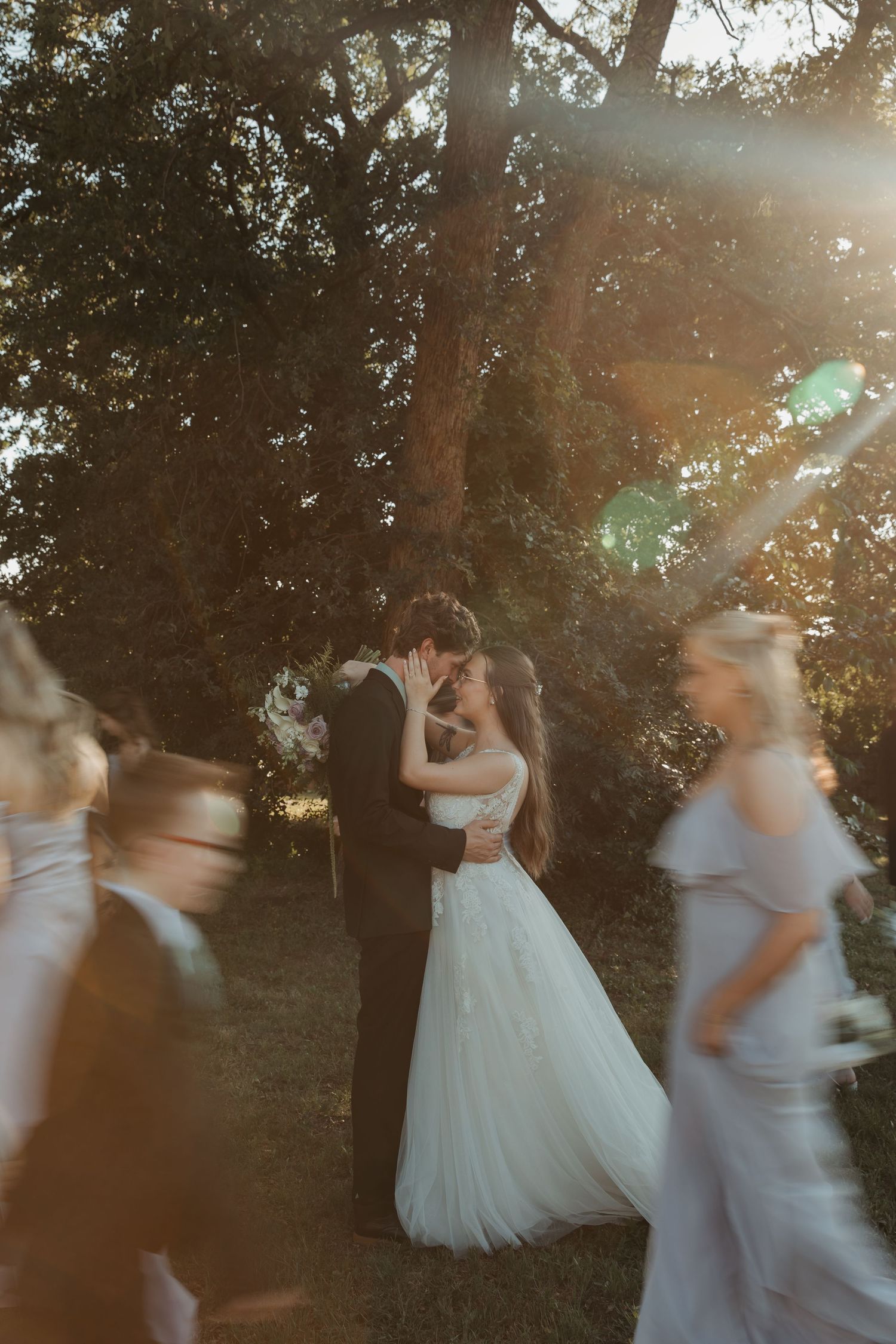 Bride and groom share a kiss outdoors in golden sunlight, surrounded by blurred wedding guests near lush green trees.