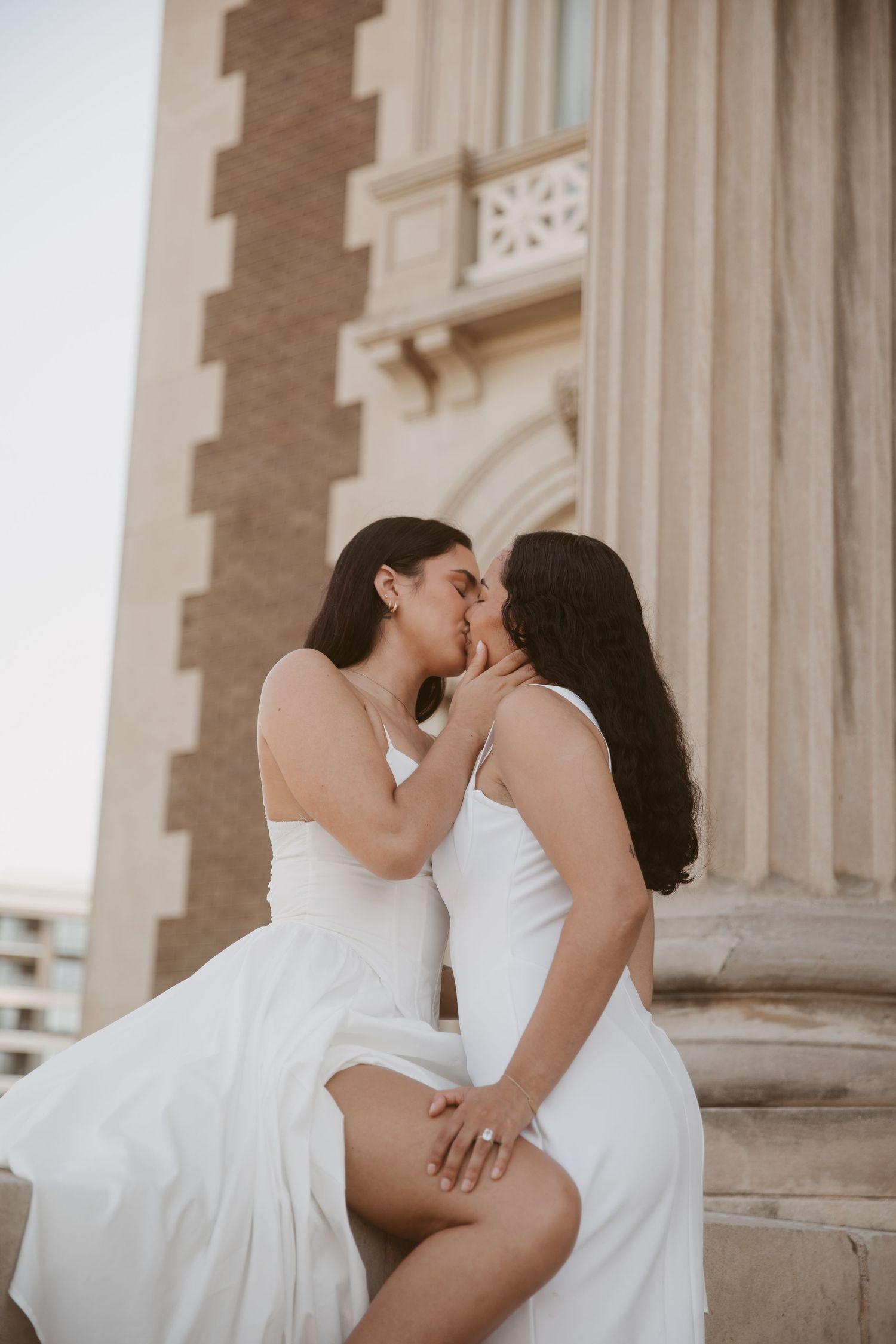 Two women in white dresses sharing a romantic kiss on the steps of a historic stone building.