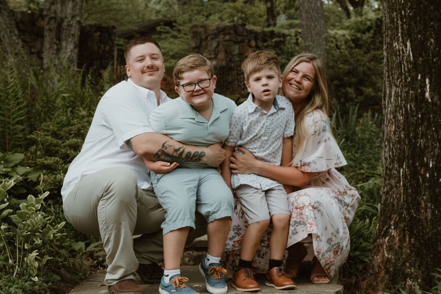 A family of four sits together outdoors on wooden steps surrounded by lush greenery in a casual summer photoshoot.