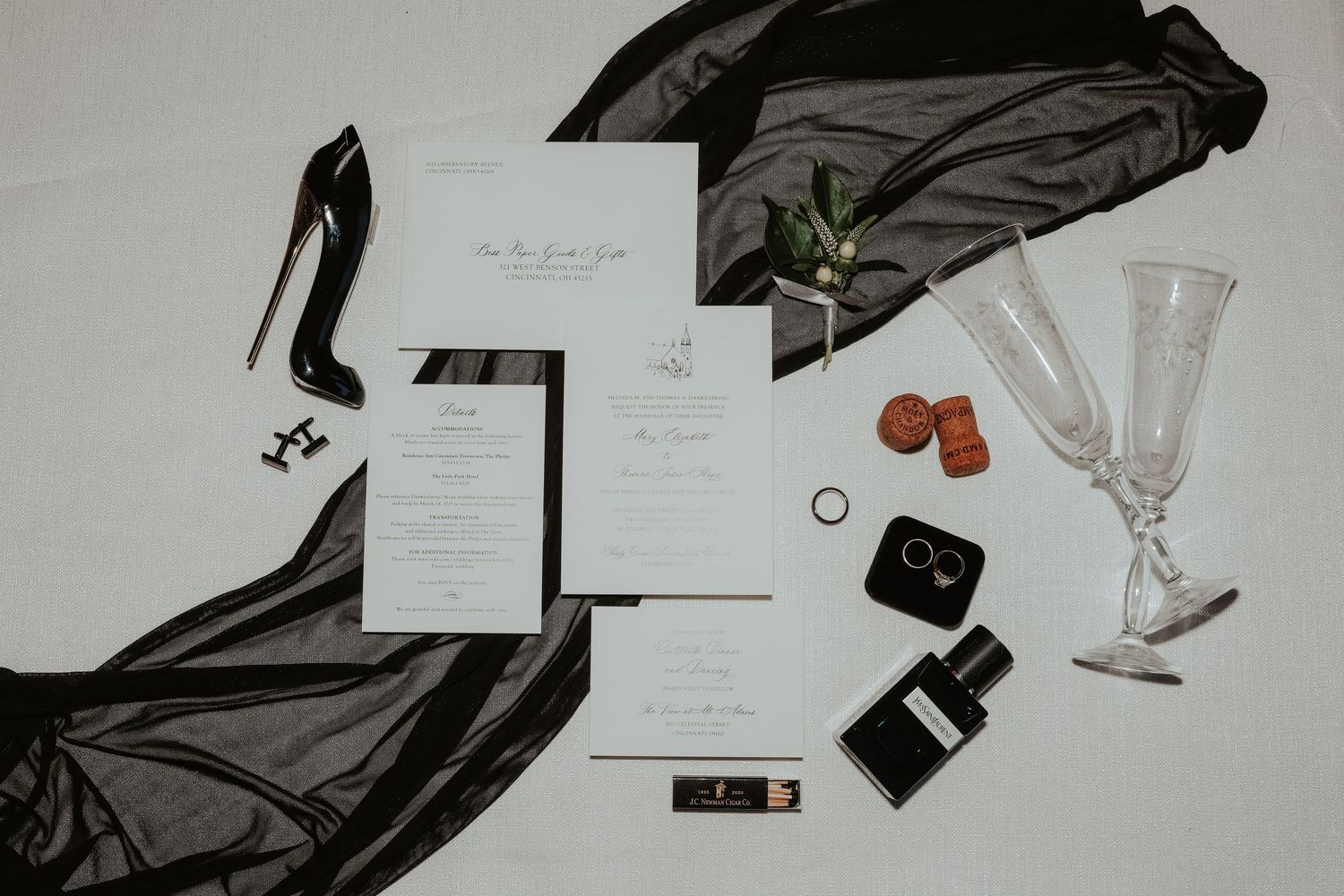 Wedding invitation flatlay with black heels, champagne flutes, and scattered accessories on white background.