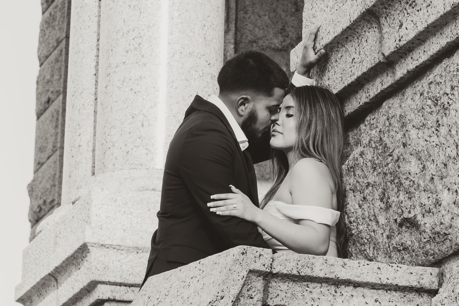 A romantic black and white portrait of a couple sharing an intimate moment against a stone pillar.