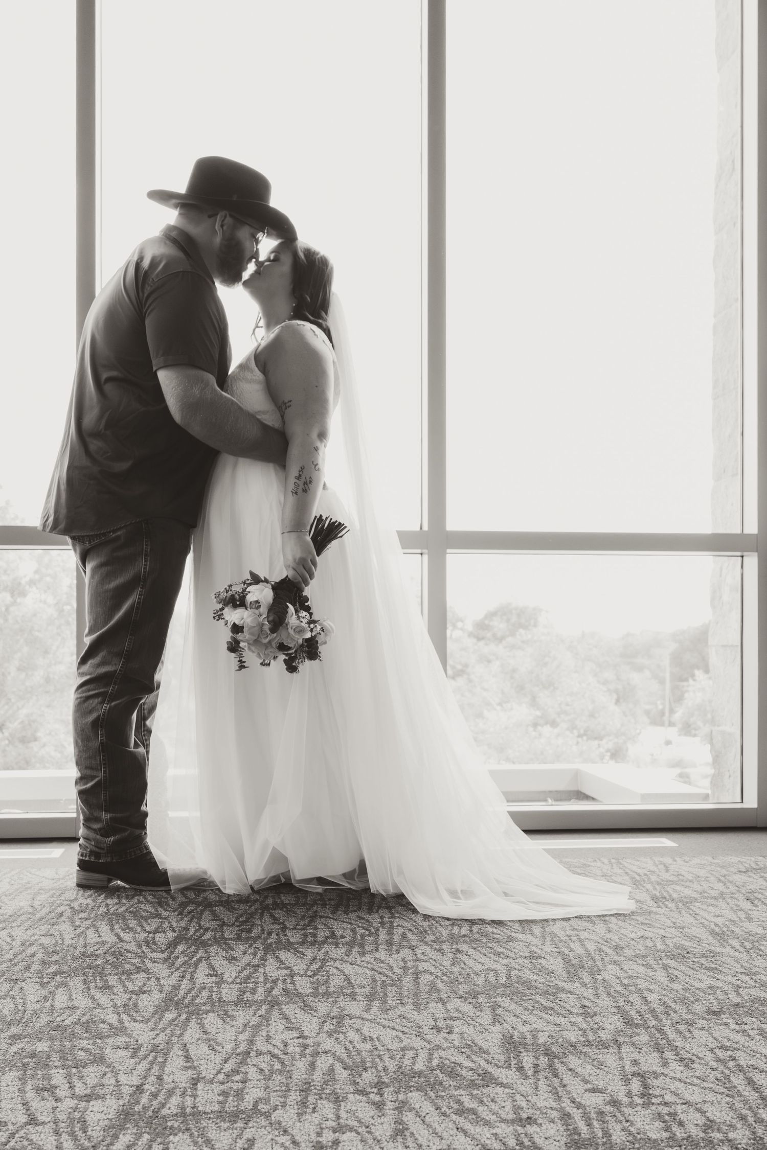 Bride and groom in western attire sharing intimate moment by window.