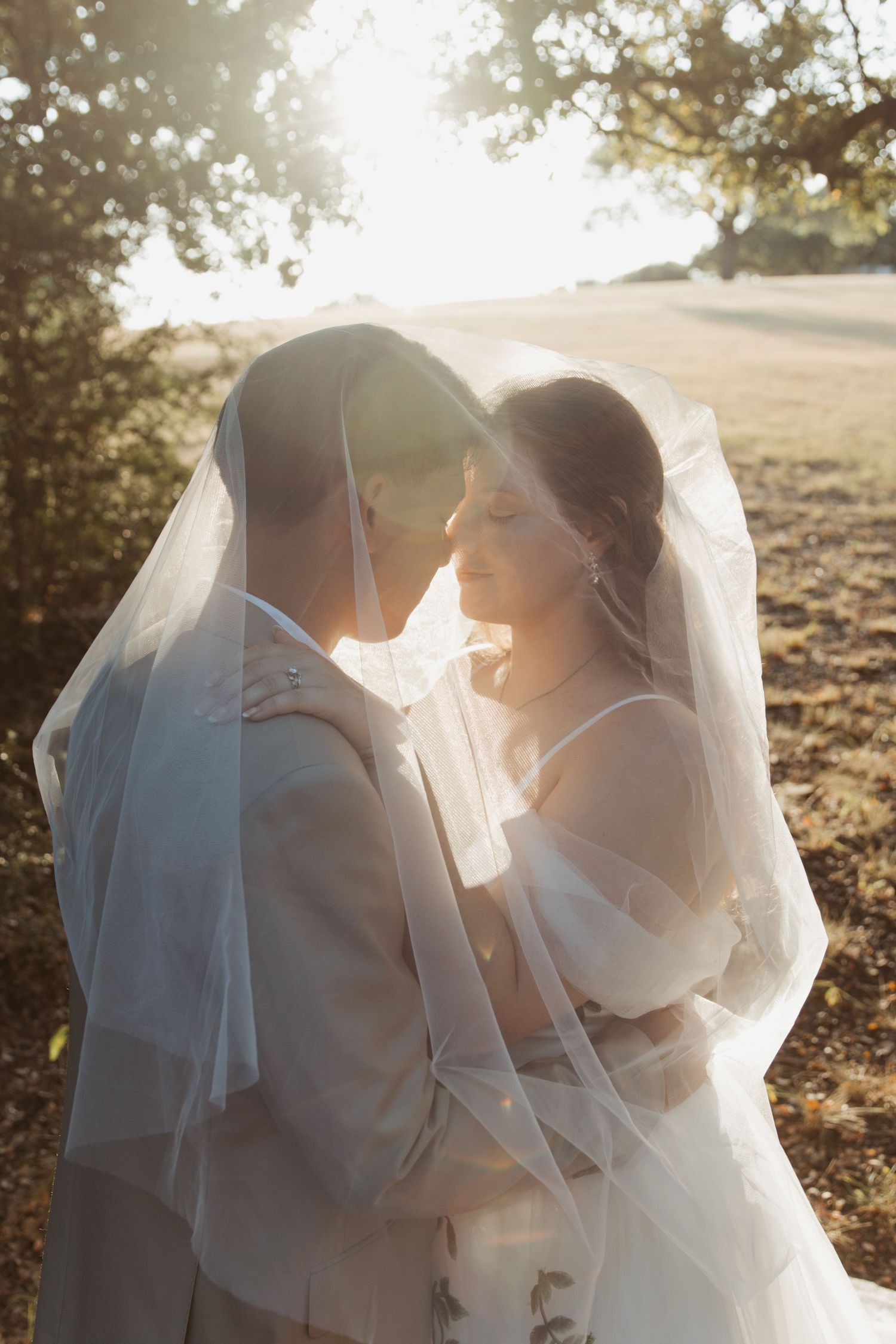 Romantic wedding couple embraces under flowing veil with golden sunlight streaming through trees at sunset.