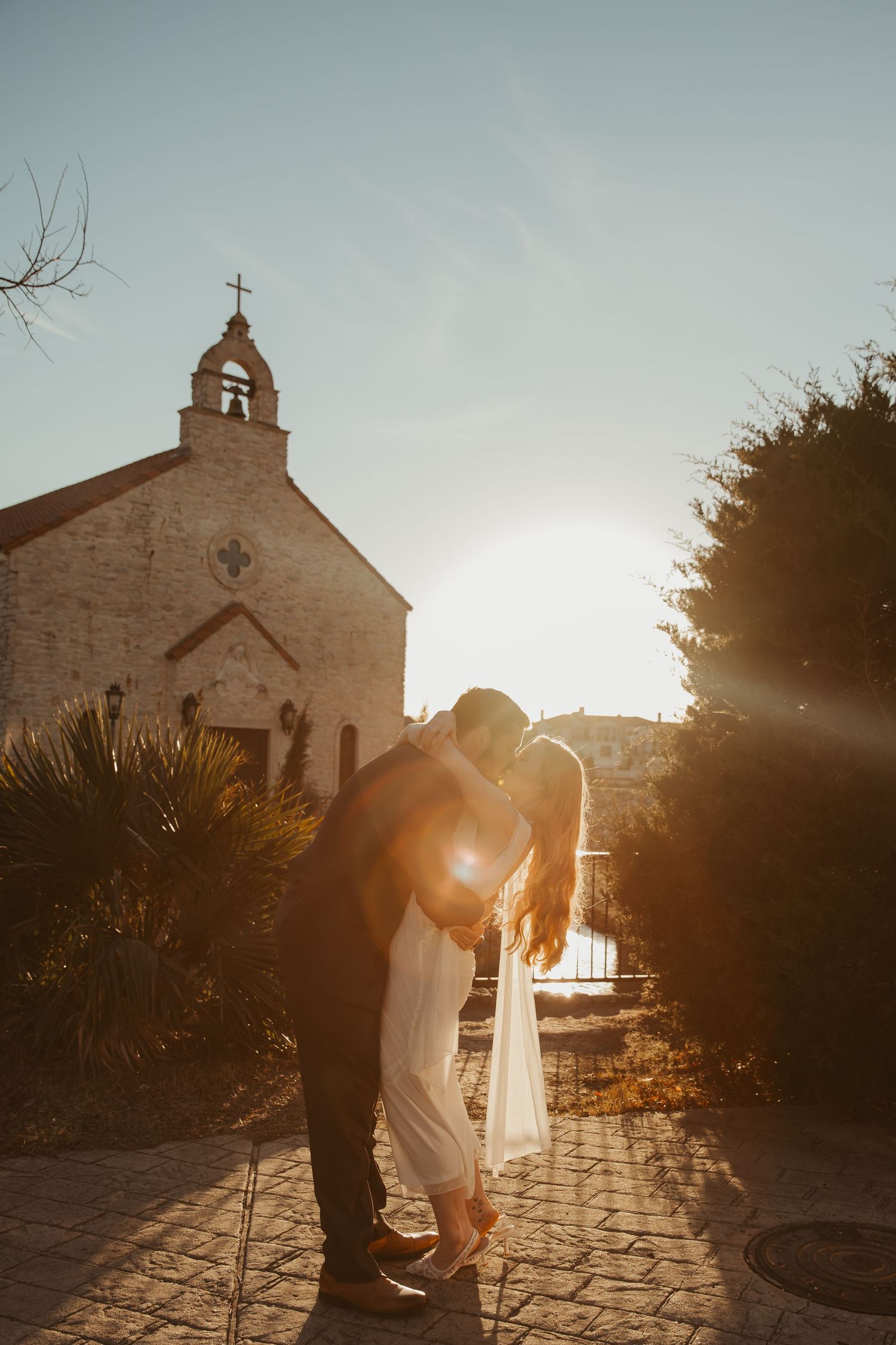 Romantic silhouettes embrace in front of a stone church during golden hour sunset with dramatic lighting.