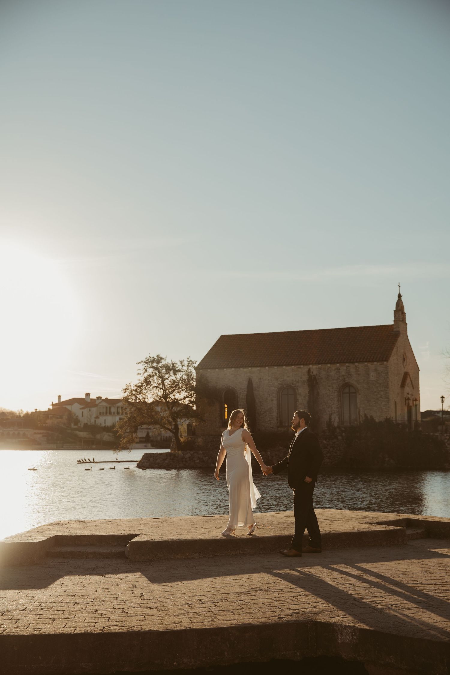 Newlyweds walk along a scenic coastal path with historic church and ocean views at golden hour.