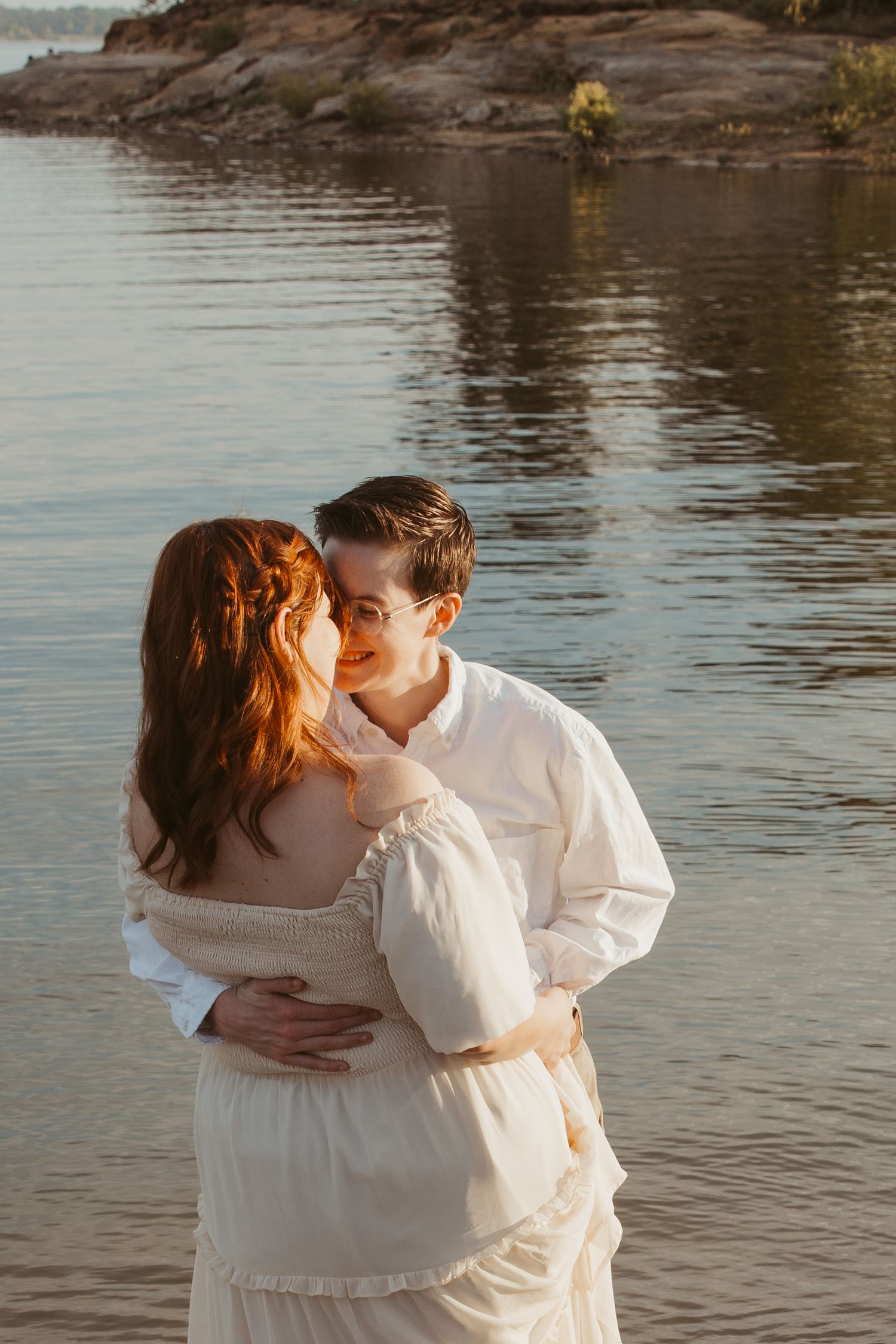 Romantic couple in white embracing by the water's edge at sunset.