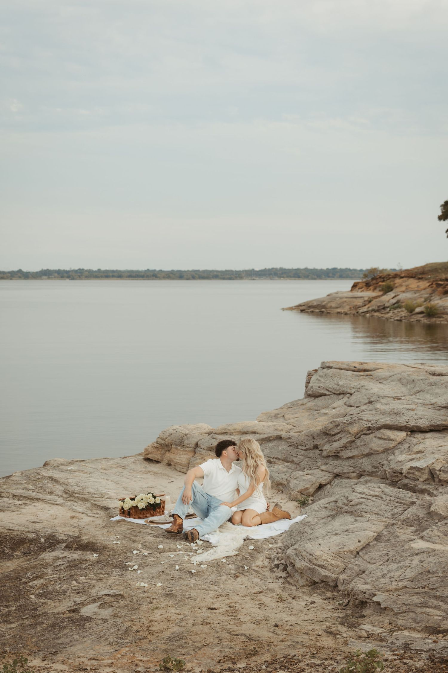 A couple in white wedding attire lies peacefully on rocky coastal cliffs overlooking a serene lake or ocean view.