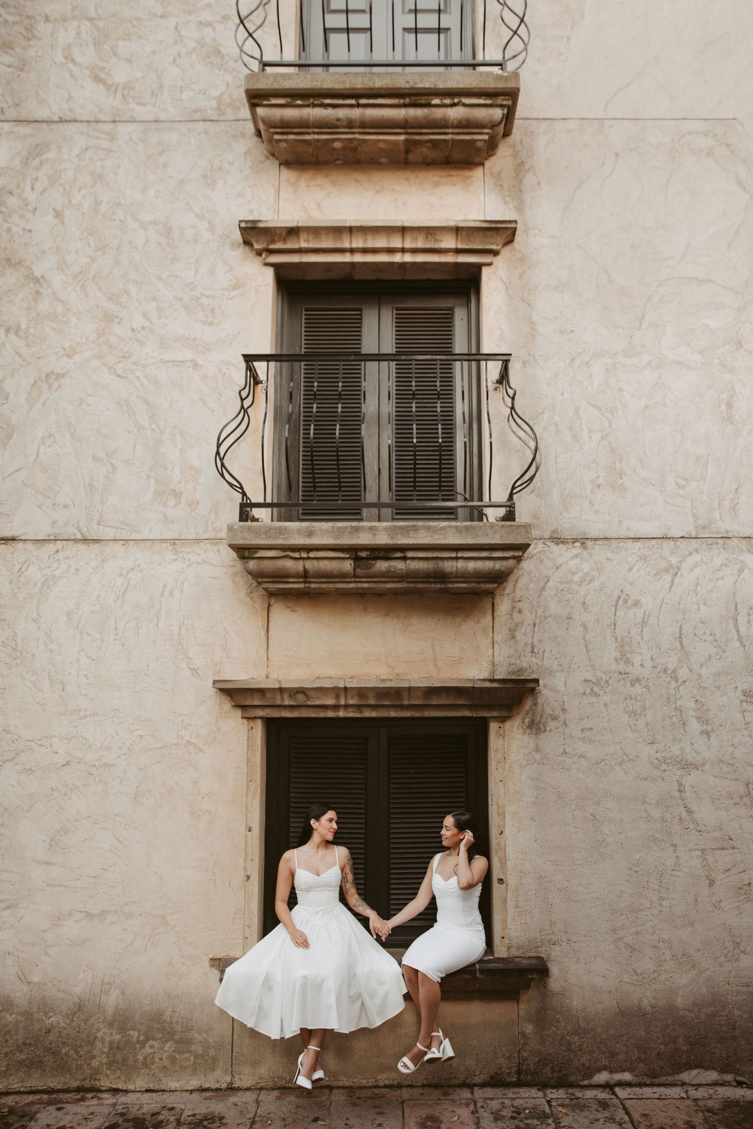 Two brides in white midi dresses hold hands outside a vintage building with balconies.