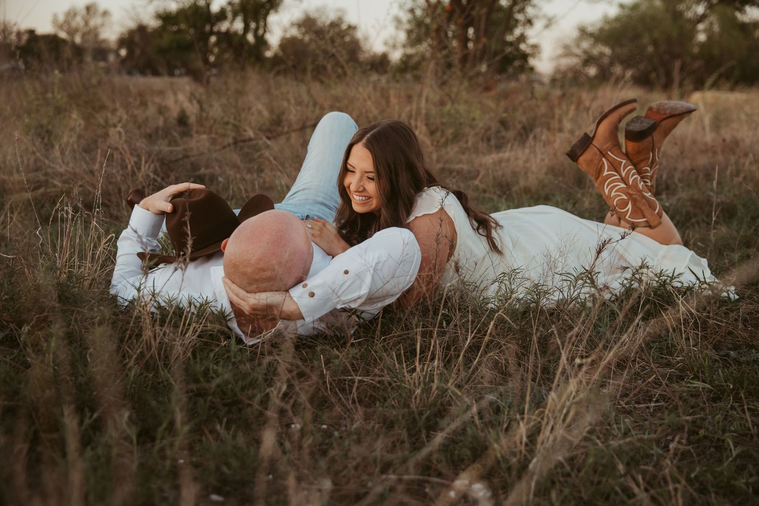 Couple laughing and lying in a golden field, woman in white dress and cowboy boots, romantic outdoor engagement photo.